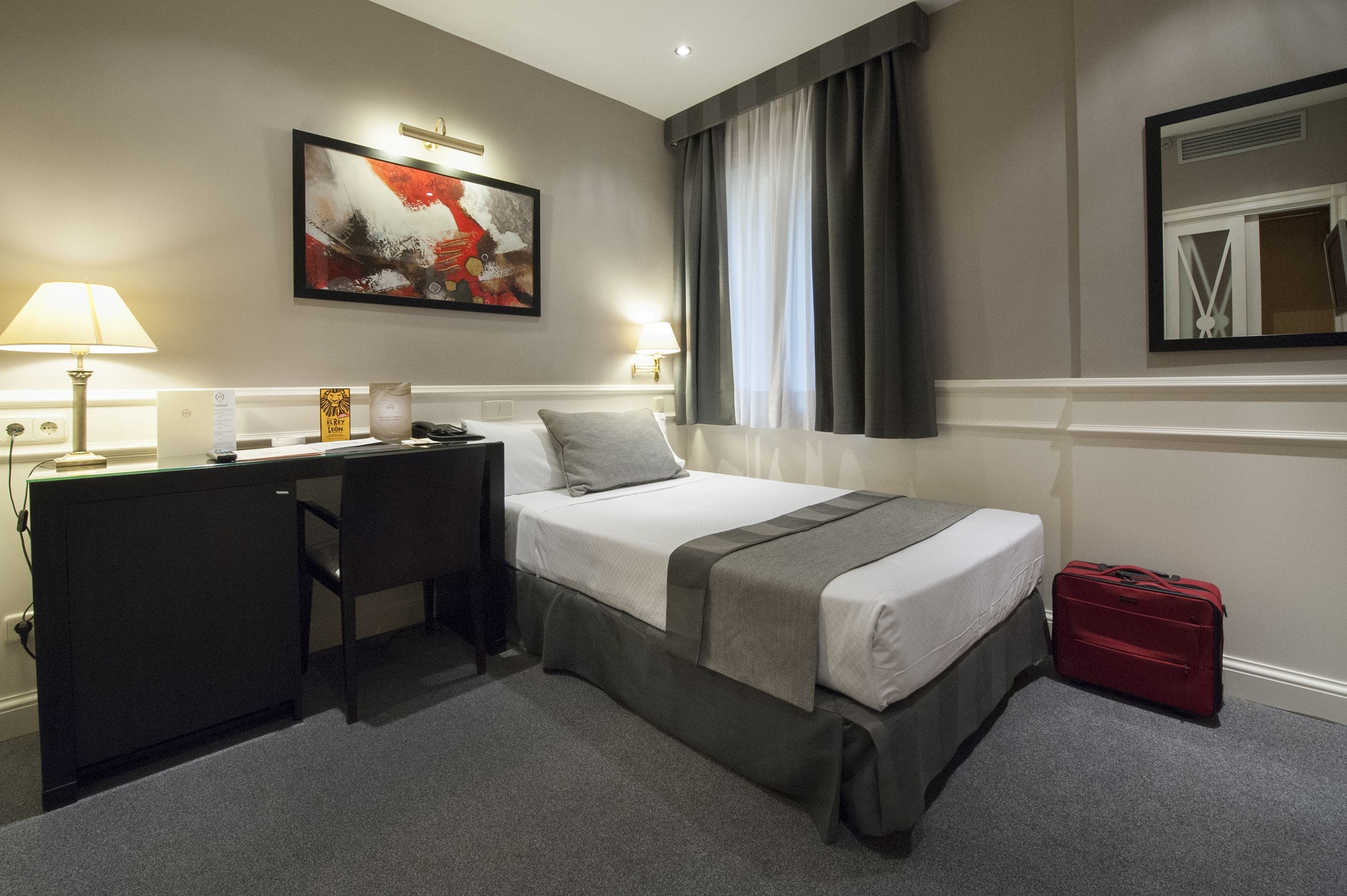 Una habitación de hotel con cama, escritorio, silla y una maleta roja.