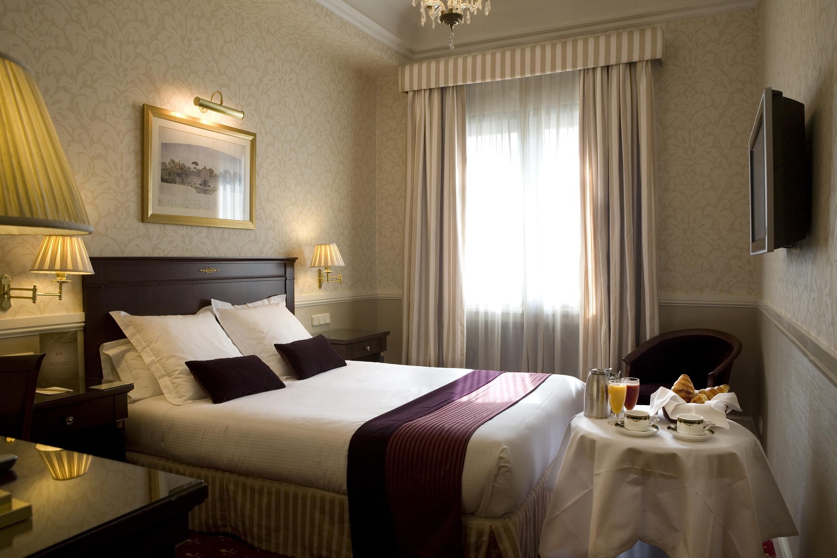 A hotel room with a large bed, a bedside table with a lamp, a small table with breakfast items, and a TV mounted on the wall.