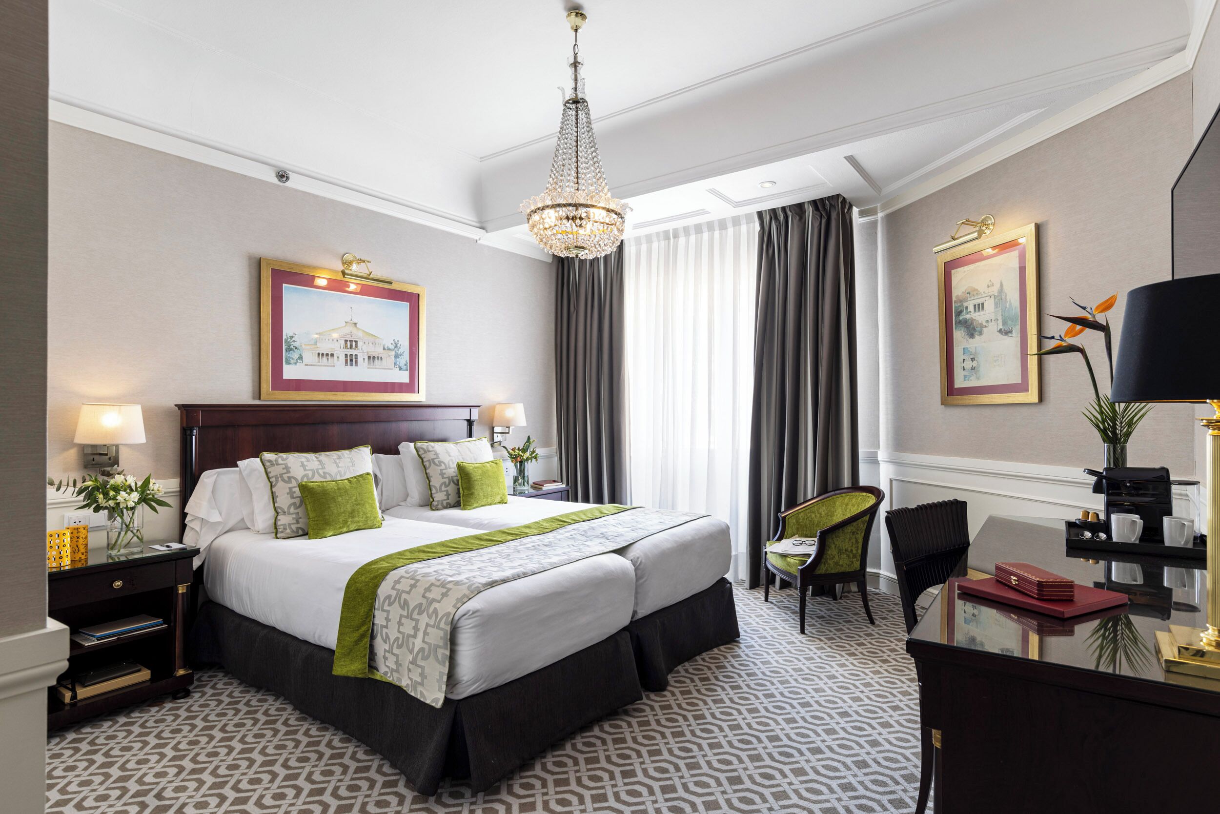 A hotel room with a large bed, a desk, a chair, a chandelier, and framed pictures on the walls.