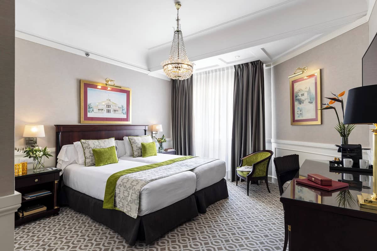 A hotel room with a large bed, a desk, a chair, a chandelier, and framed pictures on the walls.