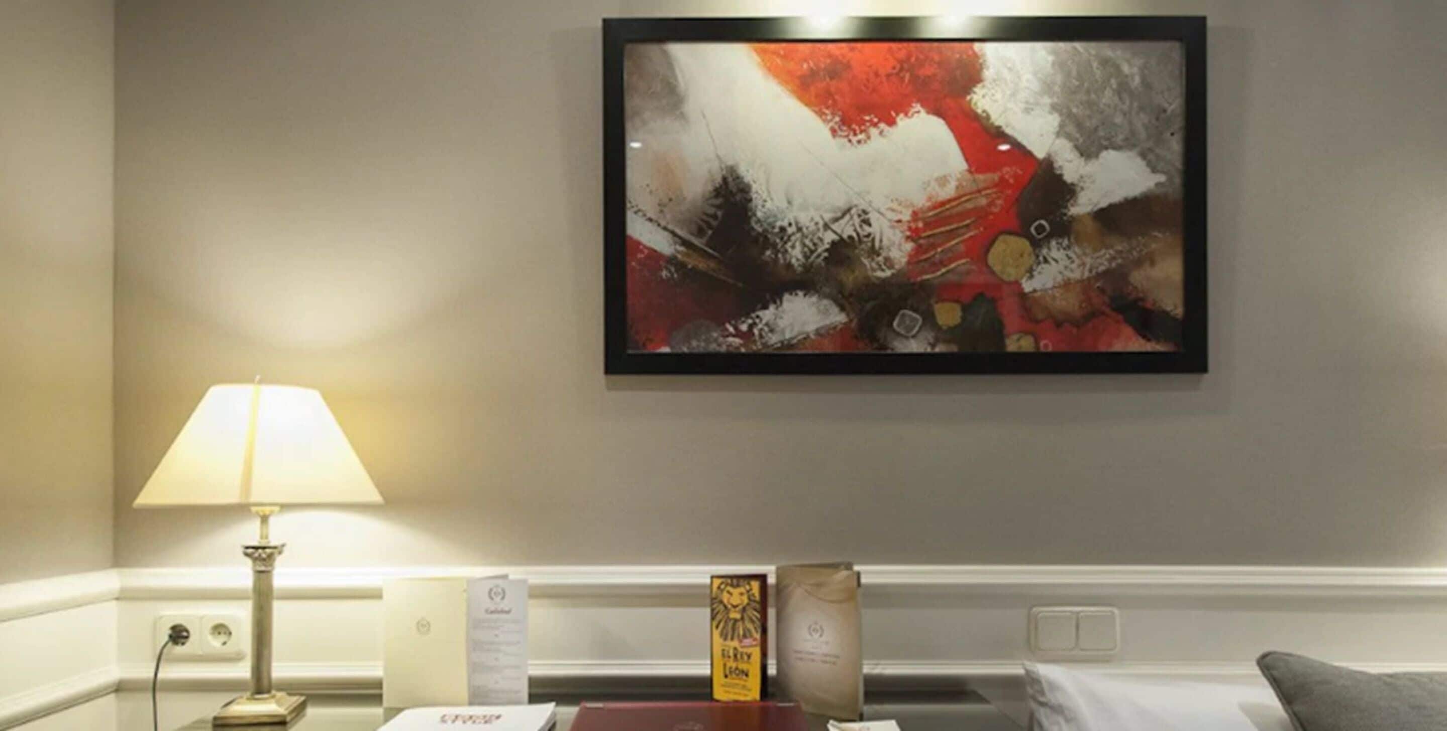 A hotel room with a modern abstract painting on the wall, a bedside lamp, a small table with a book, and a bed with a gray pillow.