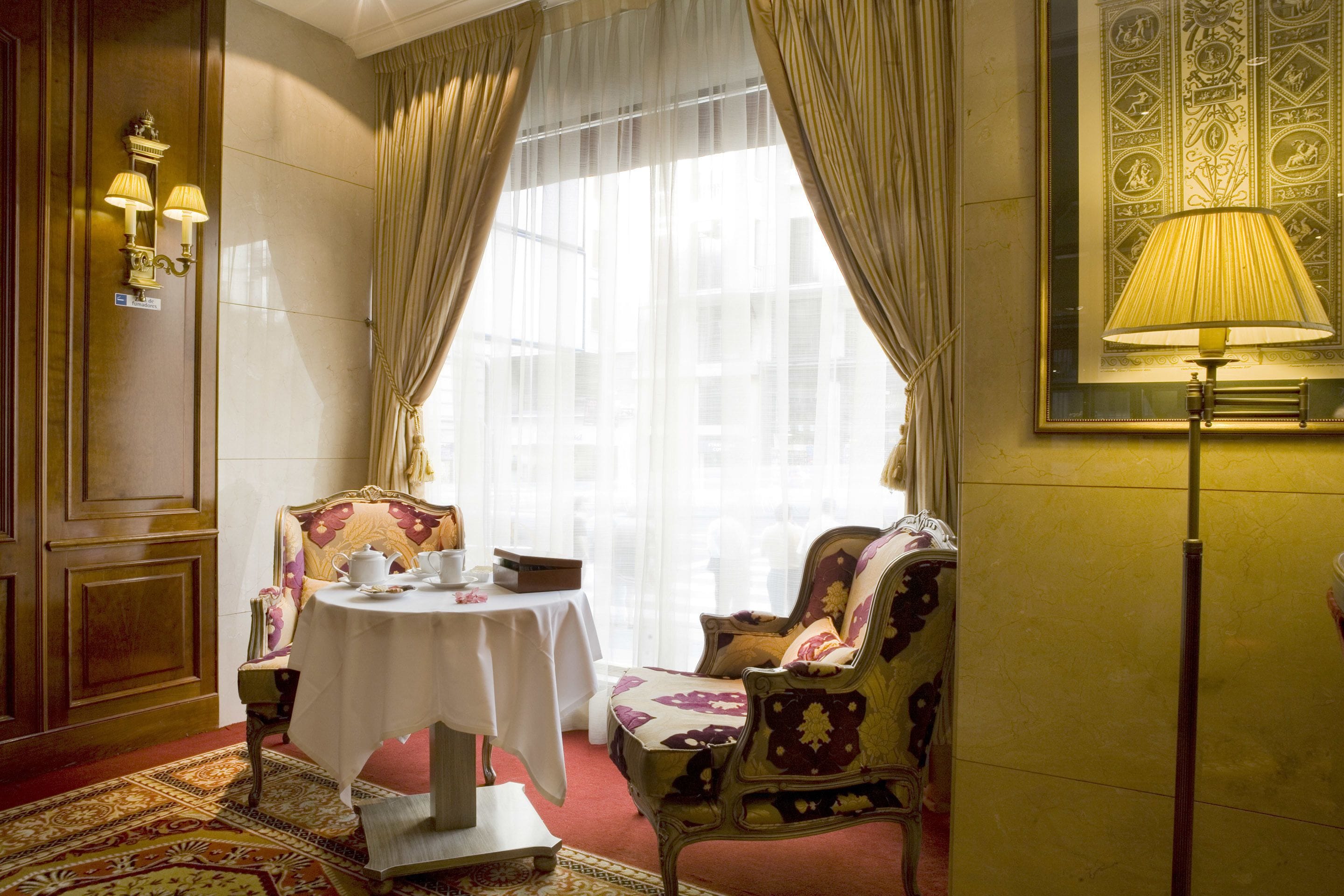 A room with a round table set for dining, two upholstered chairs, a standing lamp, and a patterned wall.