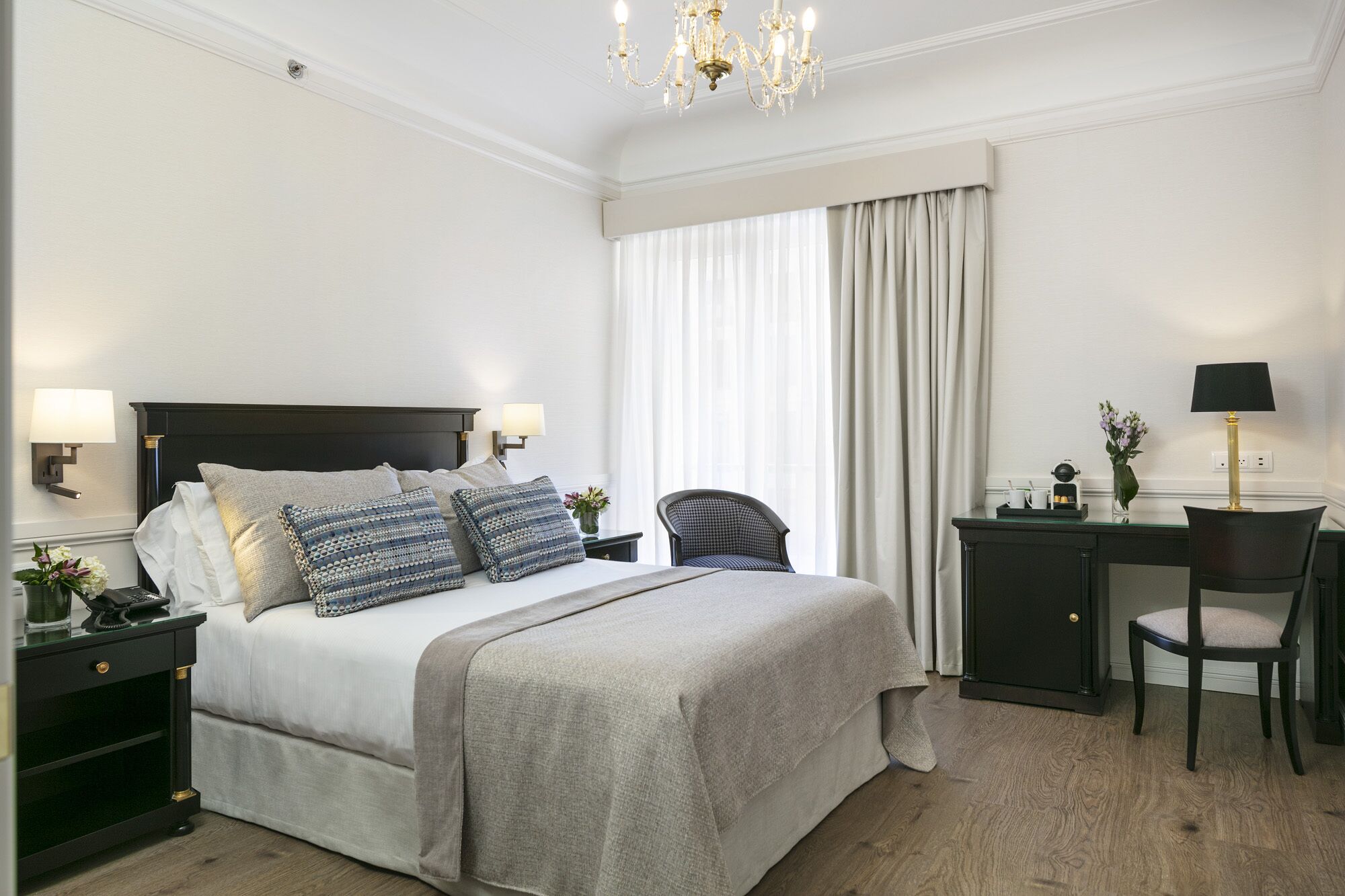 A hotel room with a large bed, two bedside tables with lamps, a desk with a chair, and a large window with curtains.