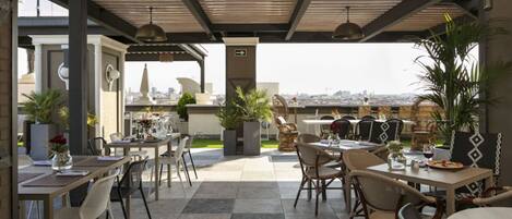 Un área de comedor al aire libre con mesas y sillas, plantas en macetas y vistas de la ciudad.