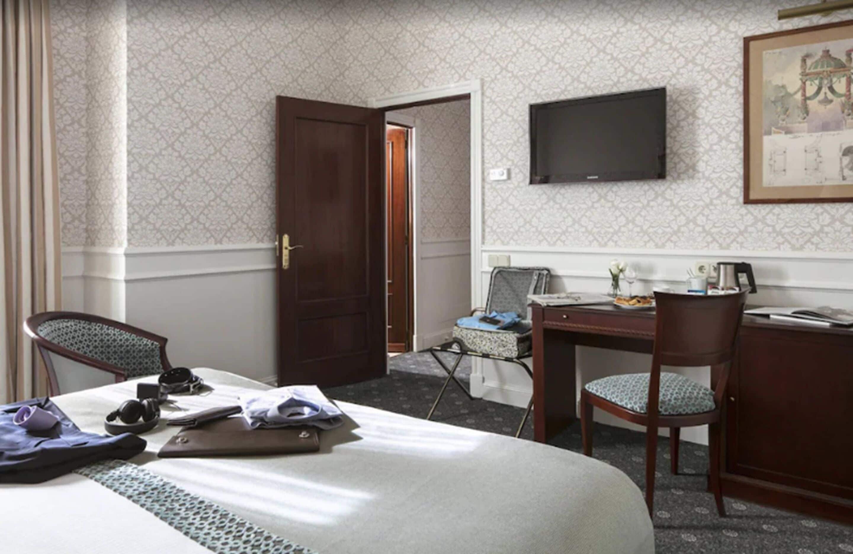 Une chambre d’hôtel comprenant un lit, deux chaises, un bureau avec un ordinateur, une télévision et une porte donnant sur une autre pièce.