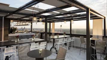 Un espacio de comedor al aire libre con mesas y sillas, un bar con vistas a la ciudad y lámparas colgantes.