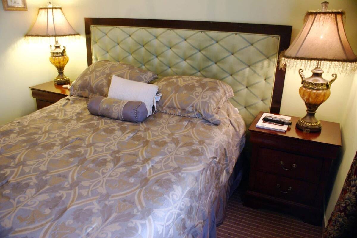Una cama bien hecha con un edredón estampado, dos almohadas y una manta doblada. Hay dos mesitas de noche con lámparas y un cajón pequeño.