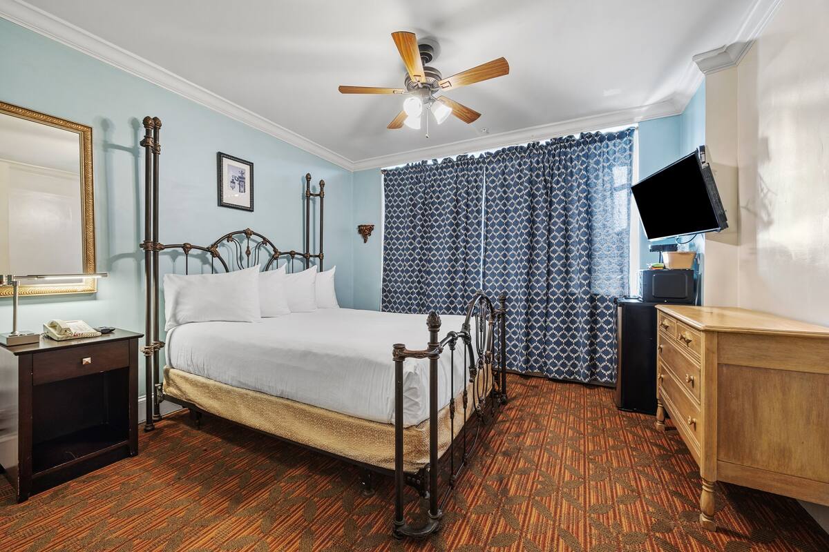Una habitación de hotel con una cama, un tocador de madera, un televisor, un ventilador de techo y cortinas estampadas.