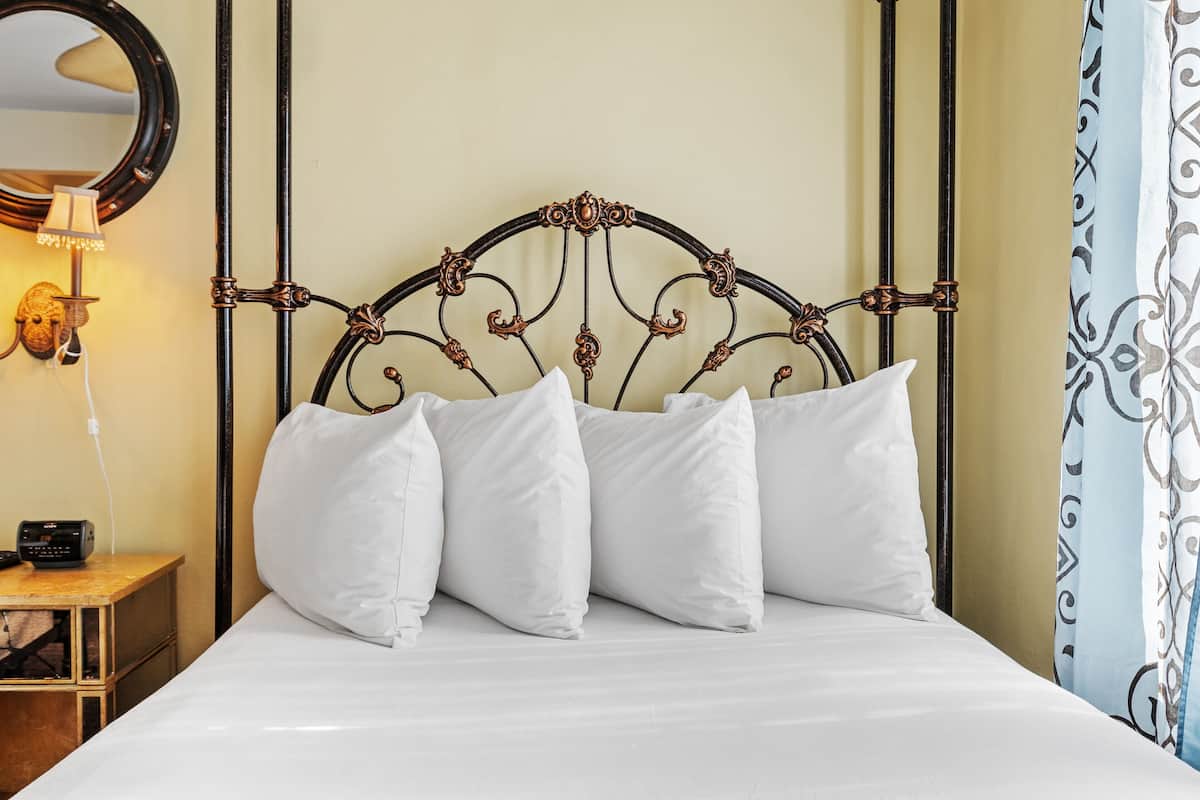 Una cama con cabecera de metal, almohadas blancas, una mesita de noche con lámpara y un espejo en la pared.