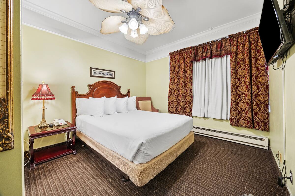 Una habitación de hotel con una cama, una mesita de noche, una lámpara, un ventilador de techo y cortinas estampadas.