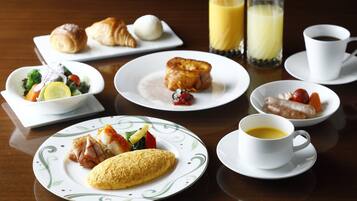 A breakfast table with croissants, eggs, a salad, a fruit plate, a plate with a rolled omelette, a cup of coffee, and two glasses of orange juice.