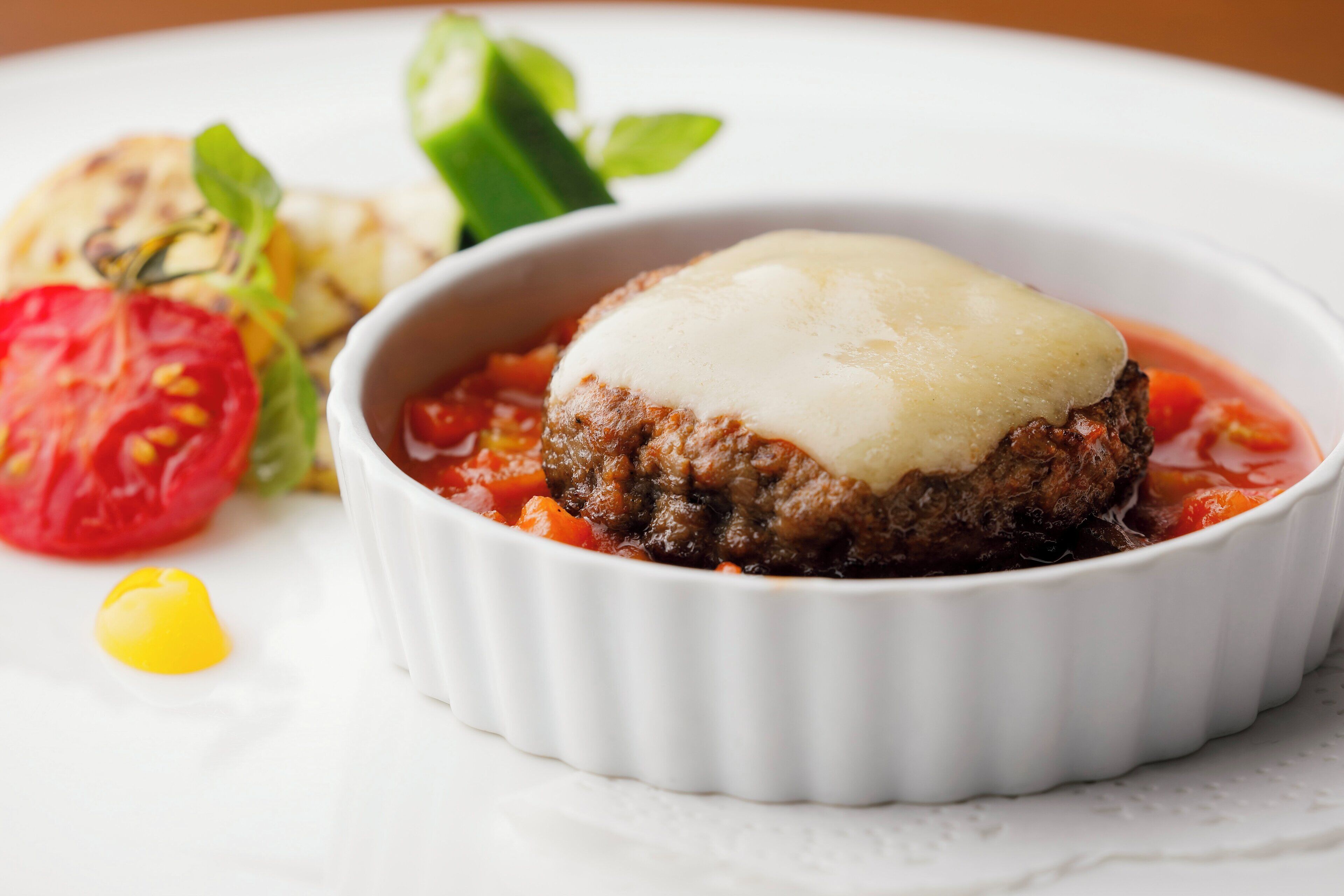 Un plato con una hamburguesa cubierta de queso en un recipiente blanco, acompañado de una ensalada y una rodaja de tomate.