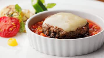 A dish with a meat patty topped with cheese in a white ramekin, accompanied by a side salad and a tomato slice.