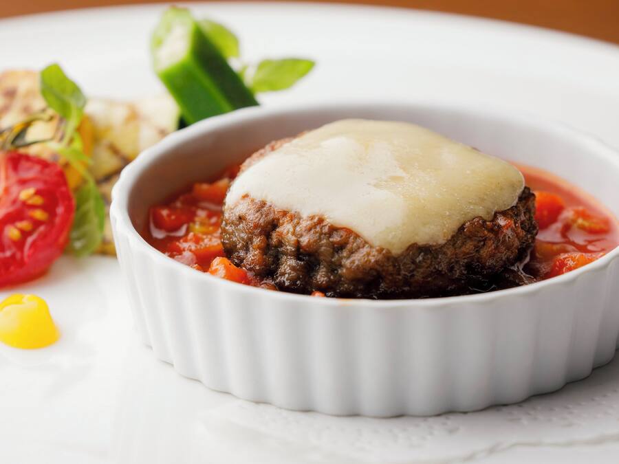 A dish with a meat patty topped with cheese in a white ramekin, accompanied by a side salad and a tomato slice.