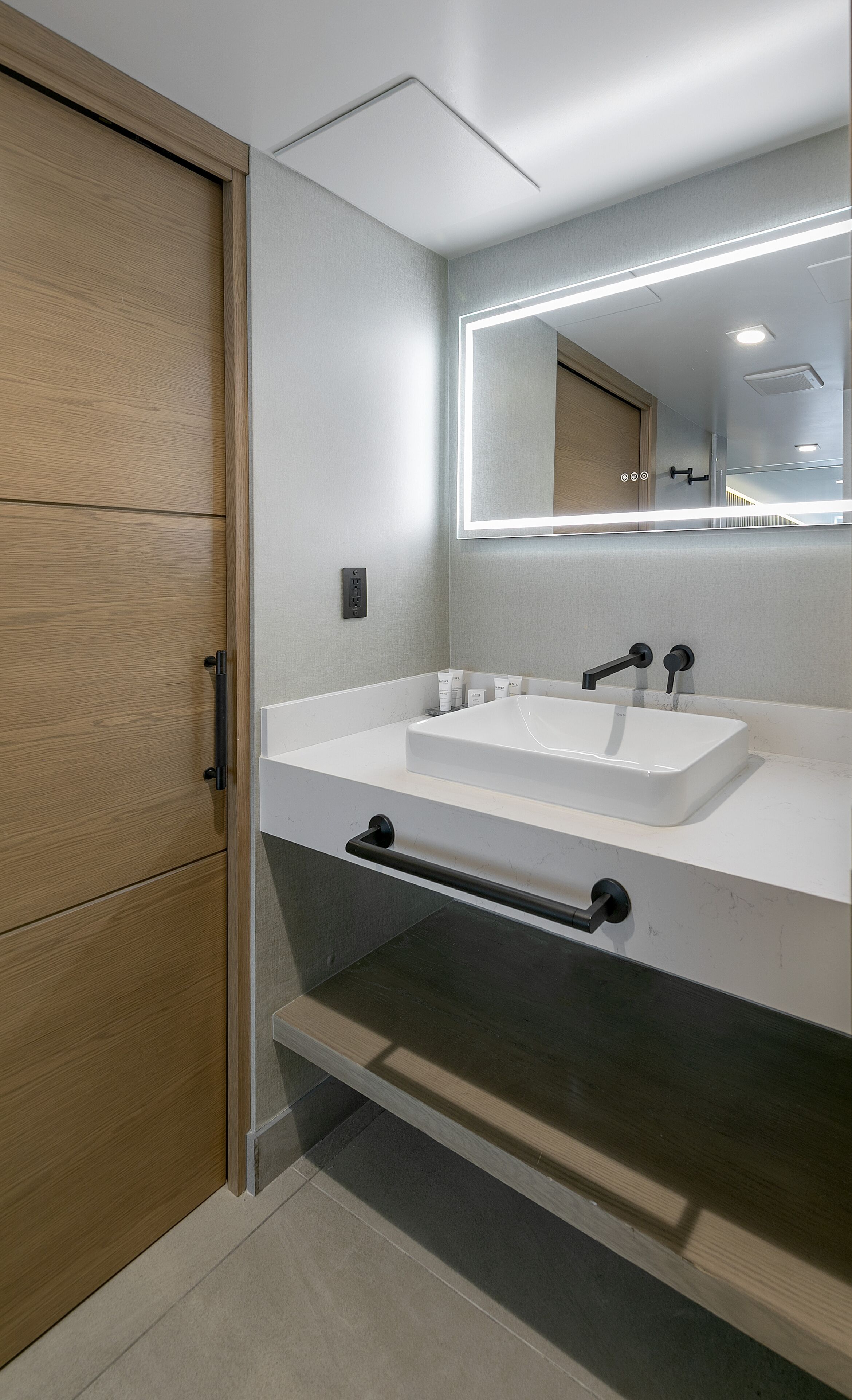 A modern bathroom with a wooden door, a white sink, and a mirror with built-in lighting.