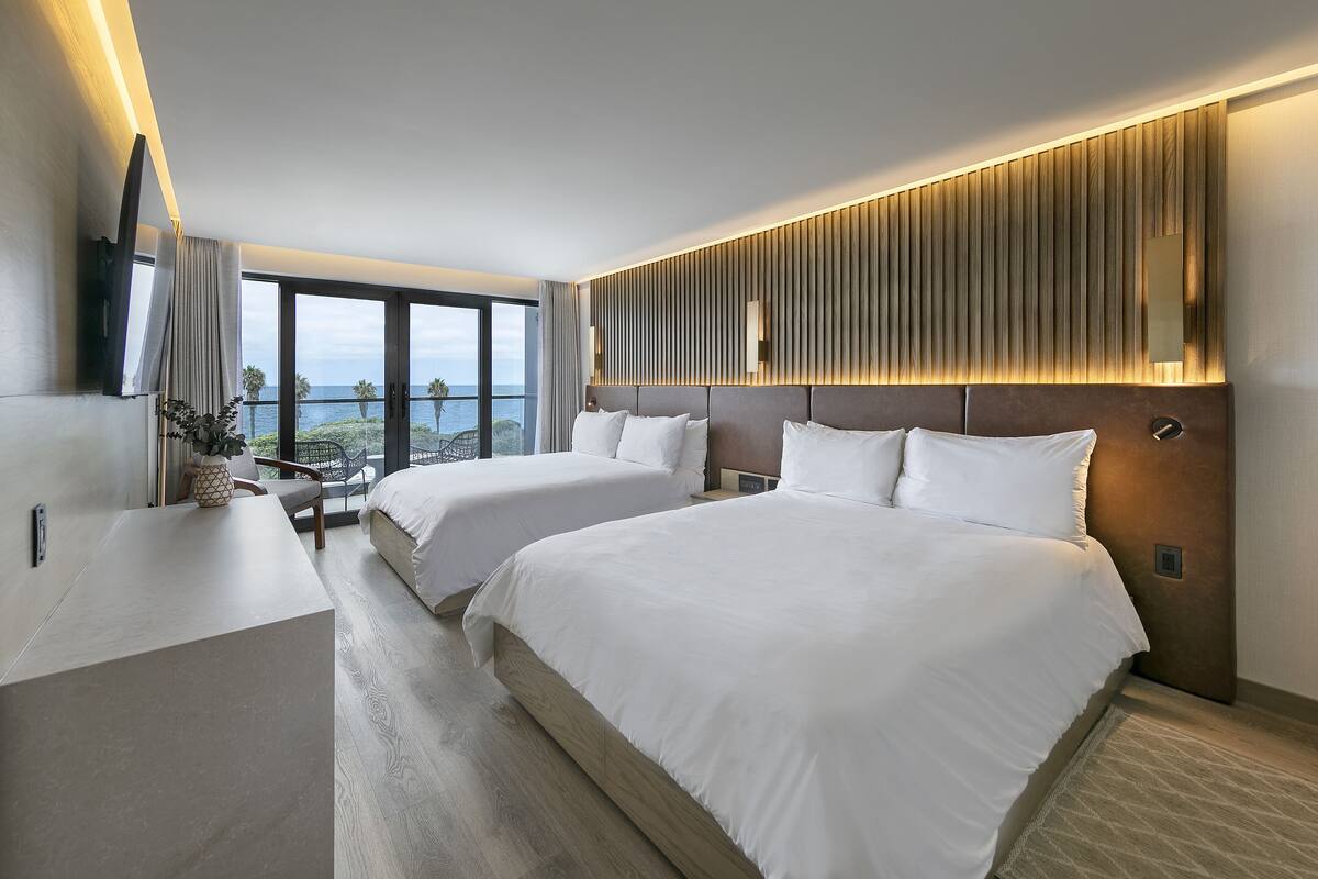A modern hotel room with two beds, a desk, and a large window offering a view of the sea.