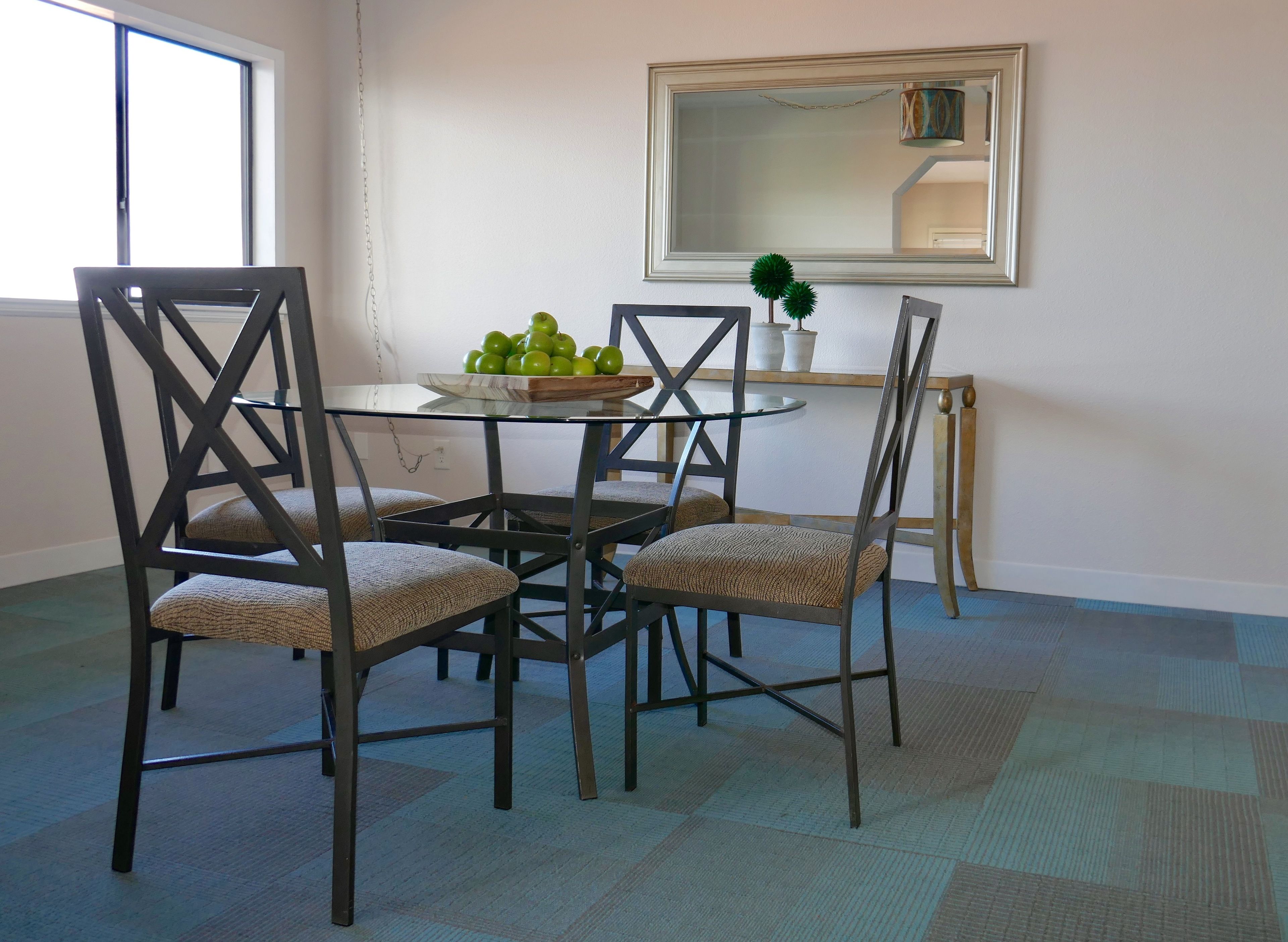 A dining area with a glass table surrounded by four chairs, a bowl of green apples, and a potted plant on a side table.