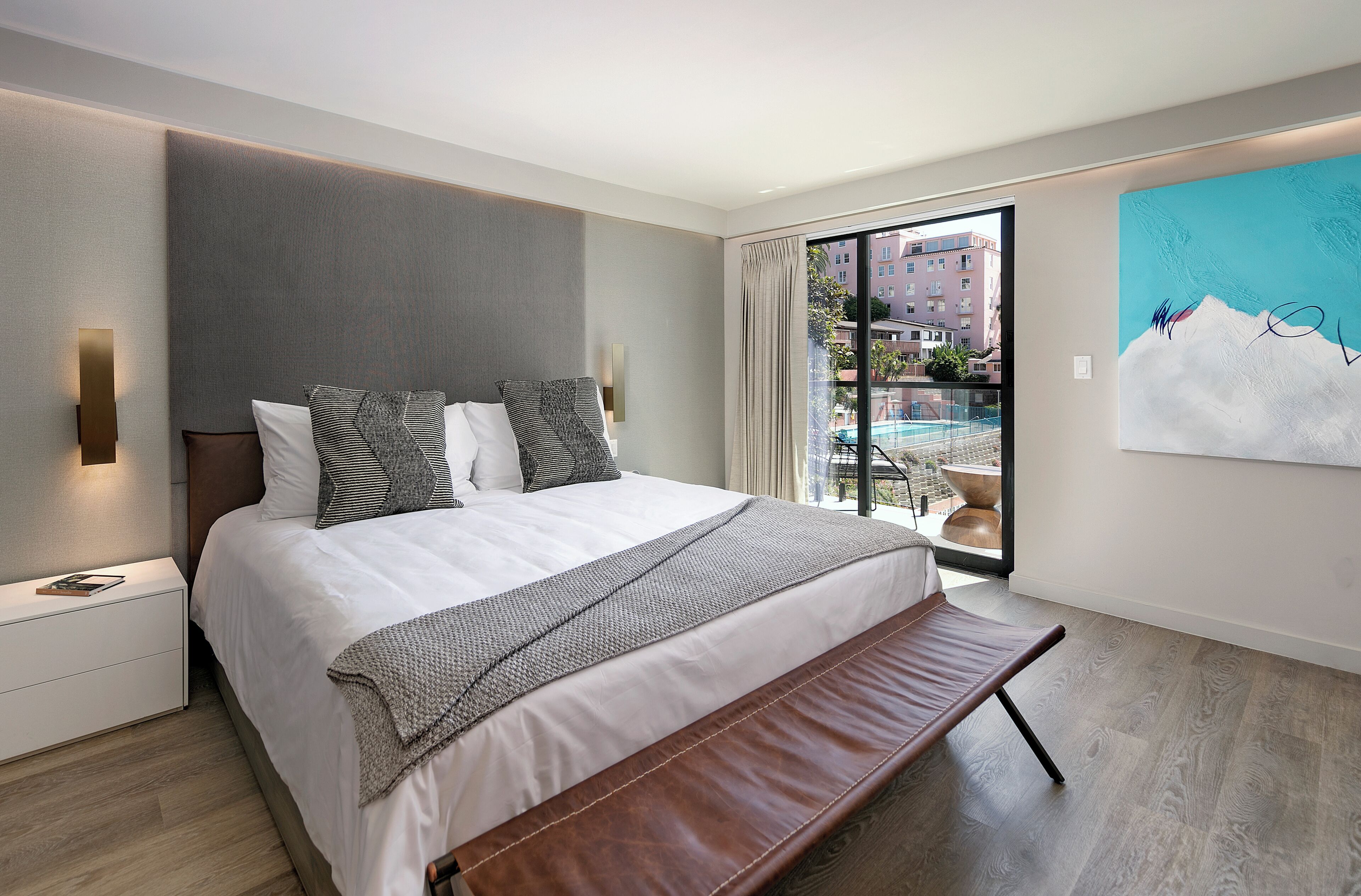 A modern bedroom with a large bed, a bench, a nightstand, and a view of a pool and buildings.