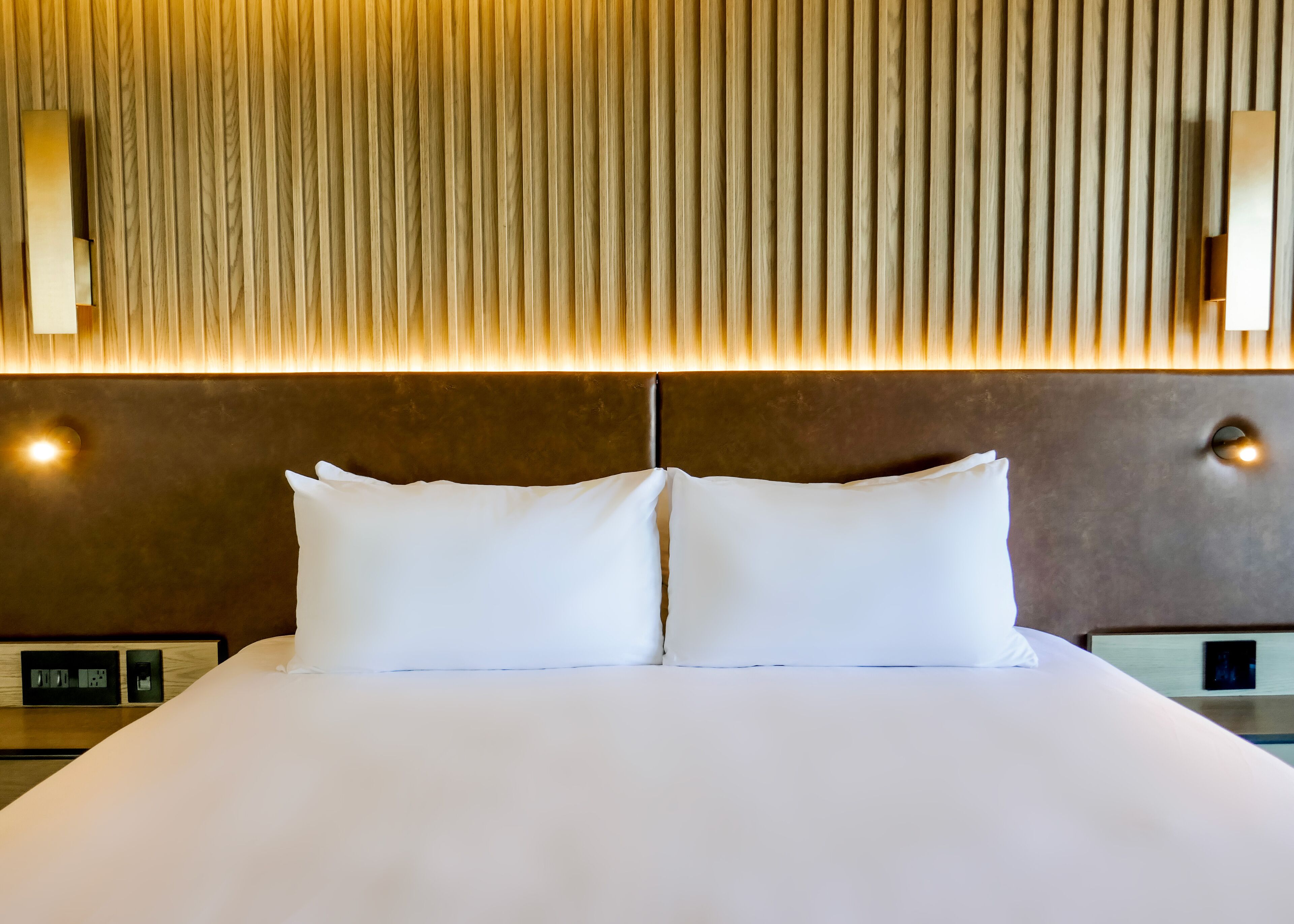 A neatly made bed with white linens and two pillows, a wooden slatted headboard, and two wall-mounted light fixtures.