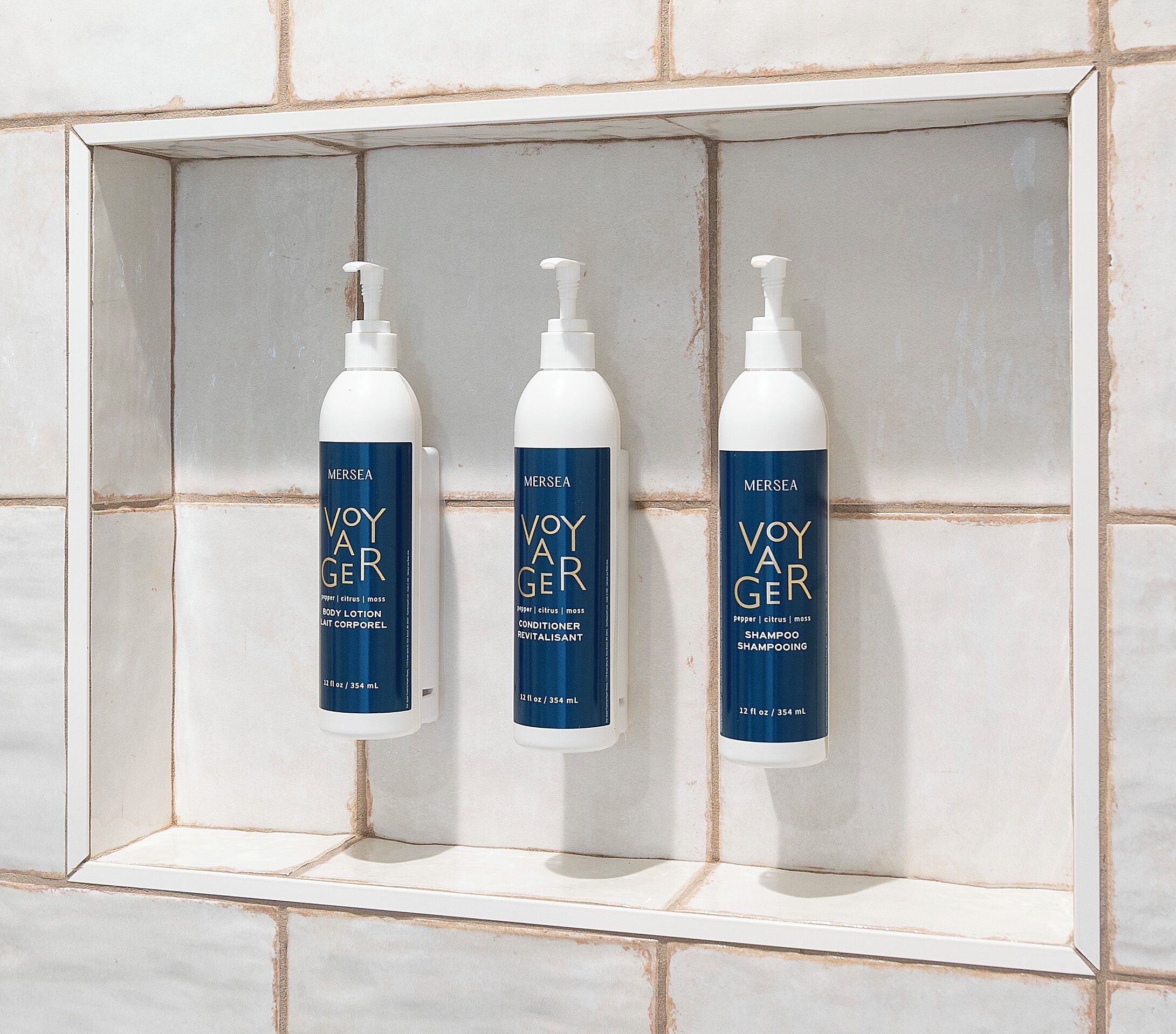 Três frascos de loção corporal, condicionador e shampoo Voyager em uma prateleira revestida de azulejos.