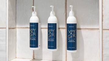 Três frascos de loção corporal, condicionador e shampoo Voyager em uma prateleira revestida de azulejos.