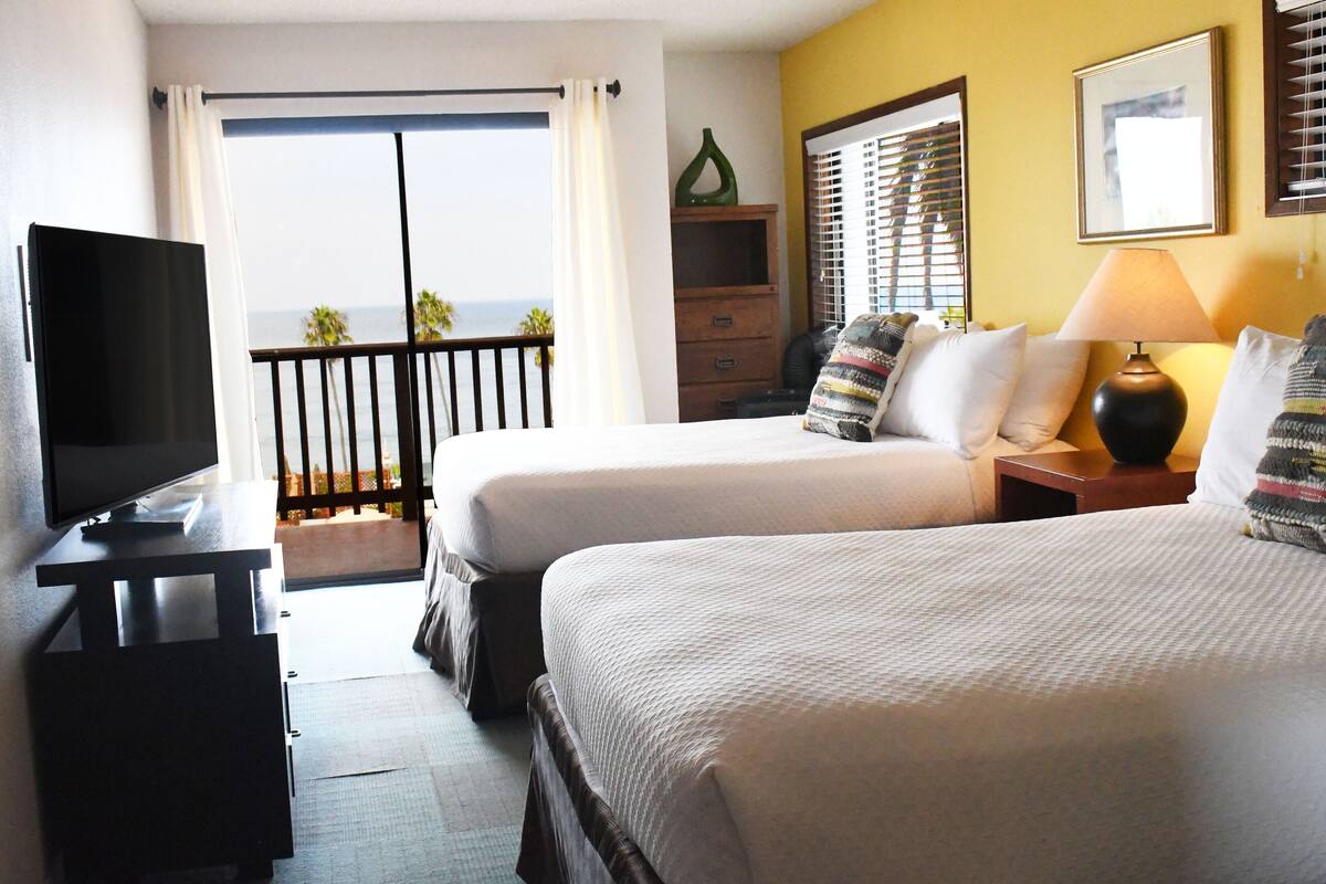 A hotel room with two beds, a TV, a lamp, a dresser, and a balcony with a view of the ocean.
