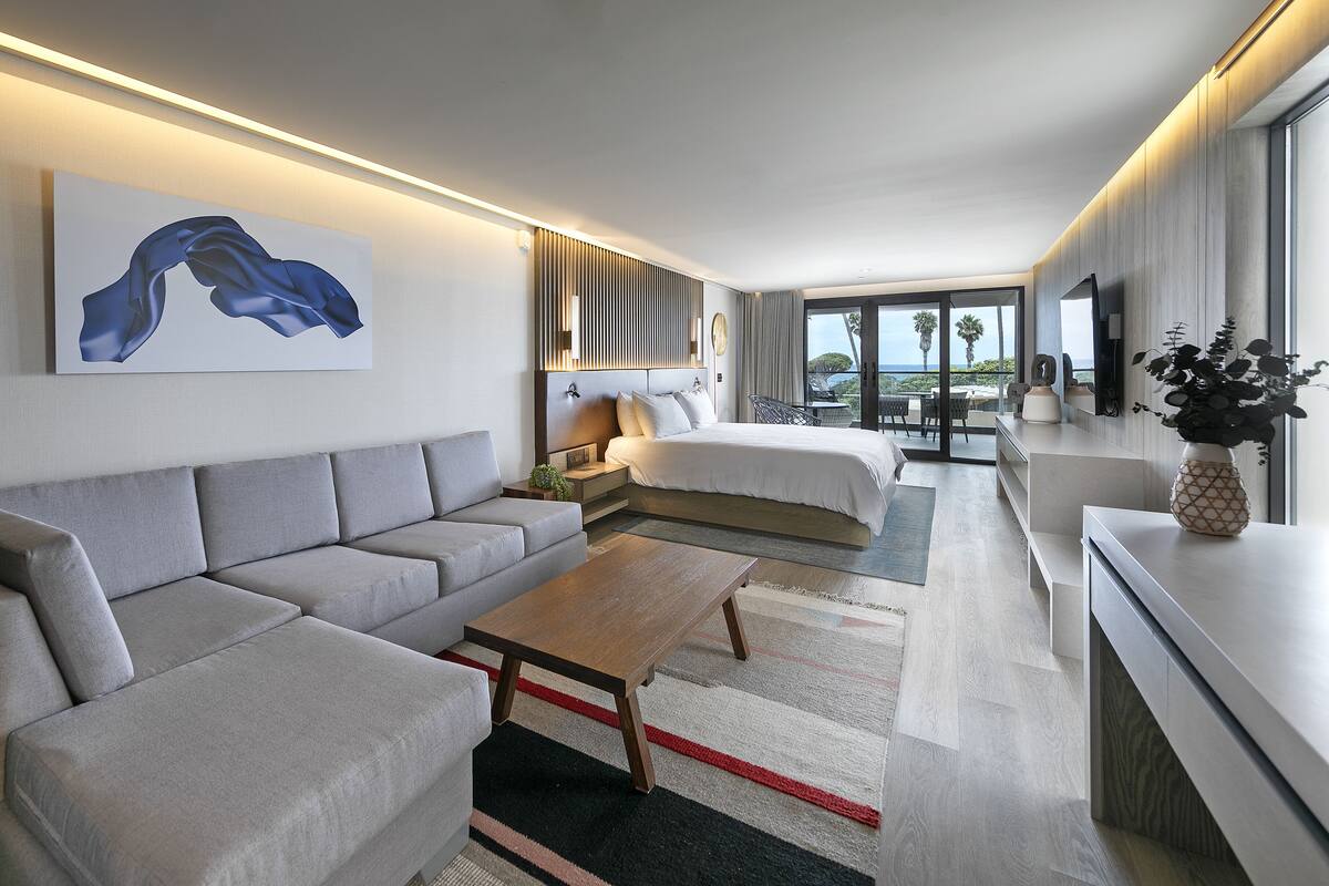 A modern hotel room with a grey sofa, a wooden coffee table, a bed with white bedding, a small side table, and a view of the ocean through large windows.