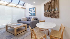 A modern living room with a grey sofa, a round white table, wicker chairs, a macrame wall hanging, and a large window with skylights. - La Jolla Cove Suites (La Jolla)