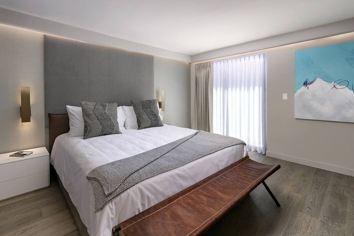 A modern bedroom with a large bed, a bench at the foot, a nightstand, and a painting on the wall.