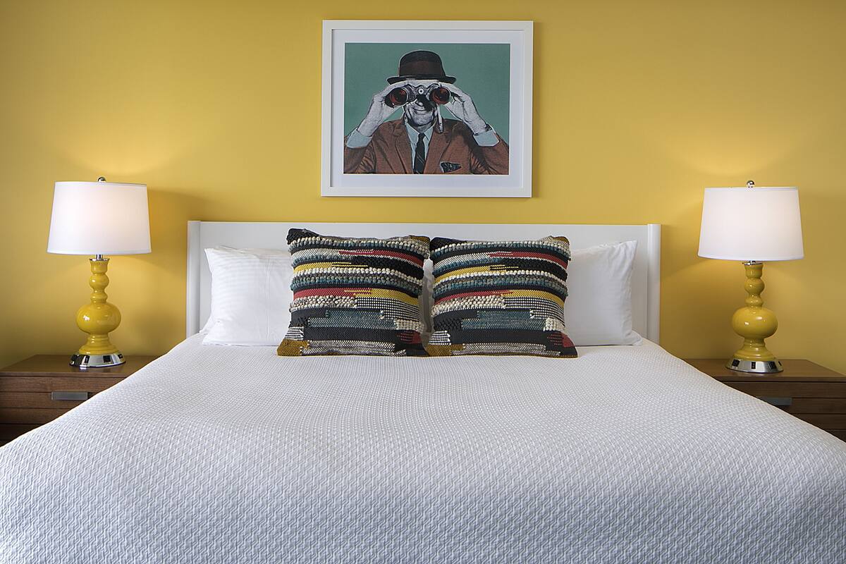 A bed with white bedding, two pillows with multicolored stripes, and a framed picture above.
