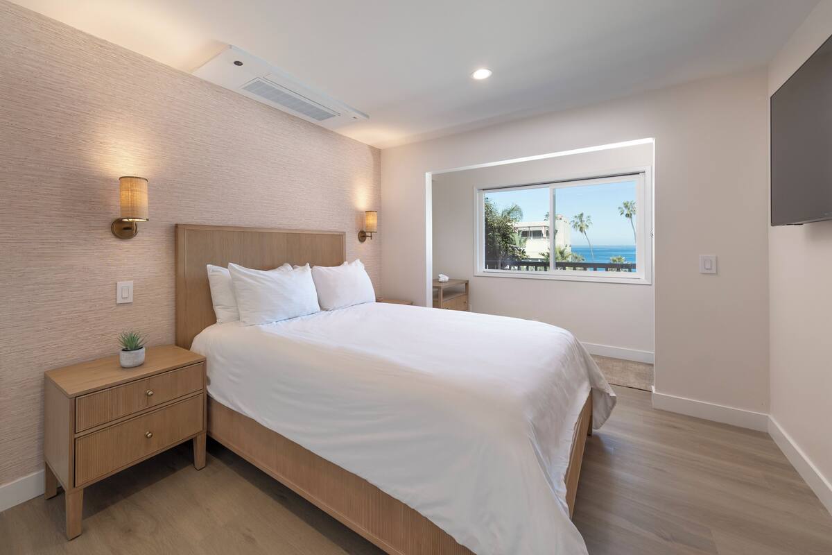 A bedroom with a bed, nightstands, a small plant, a window with a view, and a mounted TV.