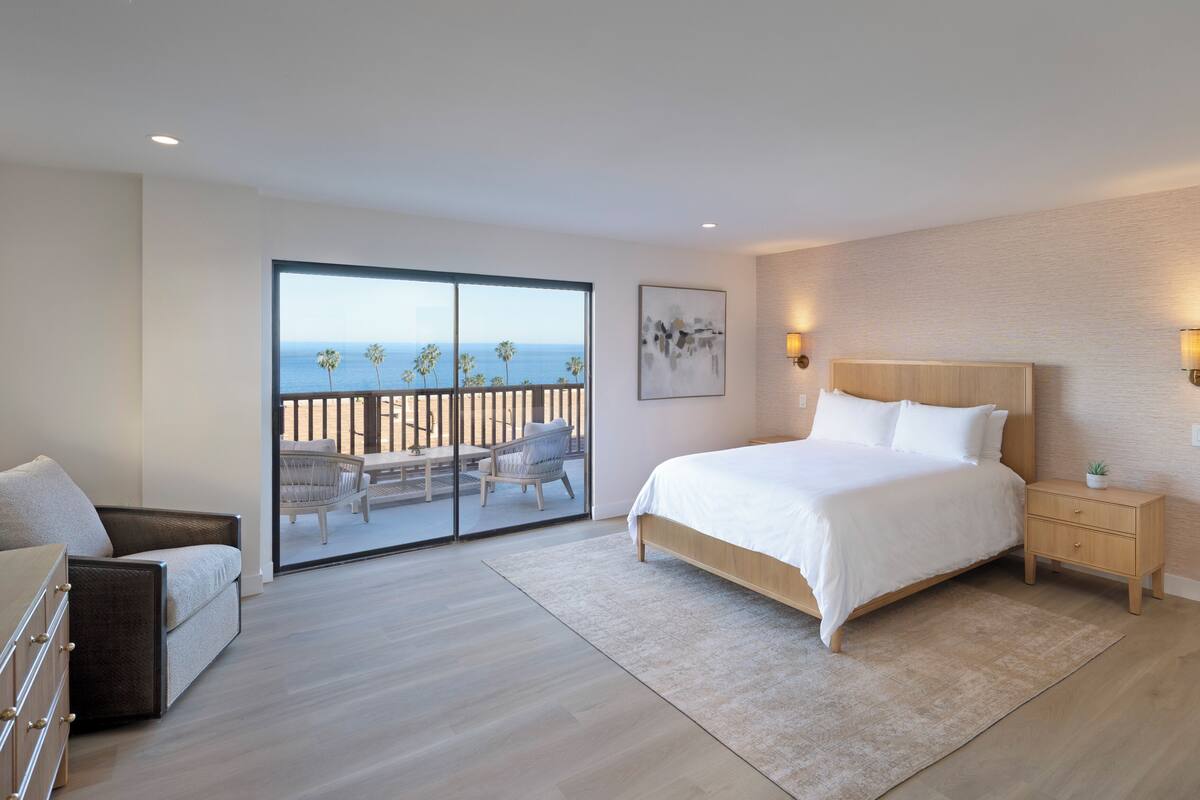 A bedroom with a large bed, a nightstand, a chair, a dresser, and a balcony with a view of the ocean.