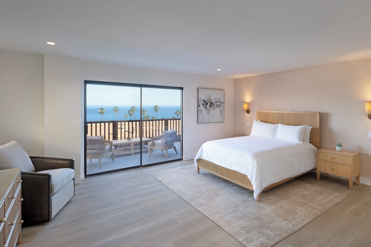 A bedroom with a large bed, a nightstand, a chair, a dresser, and a balcony with a view of the ocean.