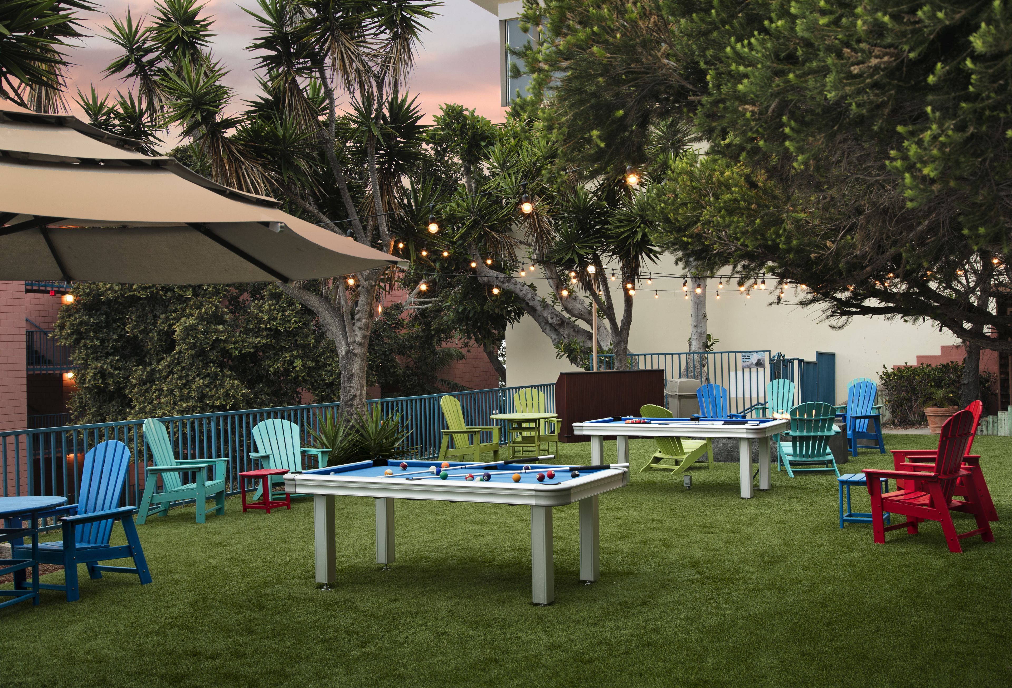 An outdoor pool area with multiple tables and chairs, surrounded by greenery and string lights.