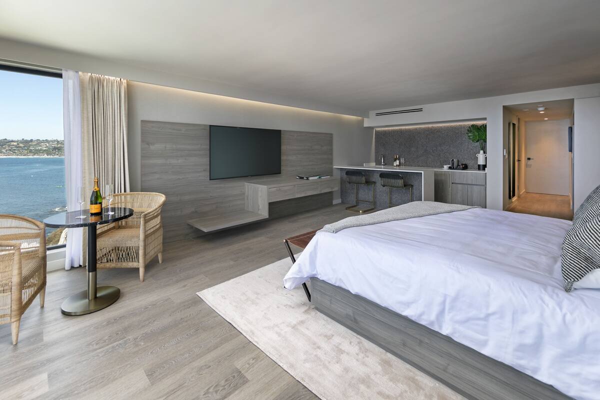 A modern hotel room with a large bed, a seating area with a table, a TV mounted on the wall, and a view of the sea.