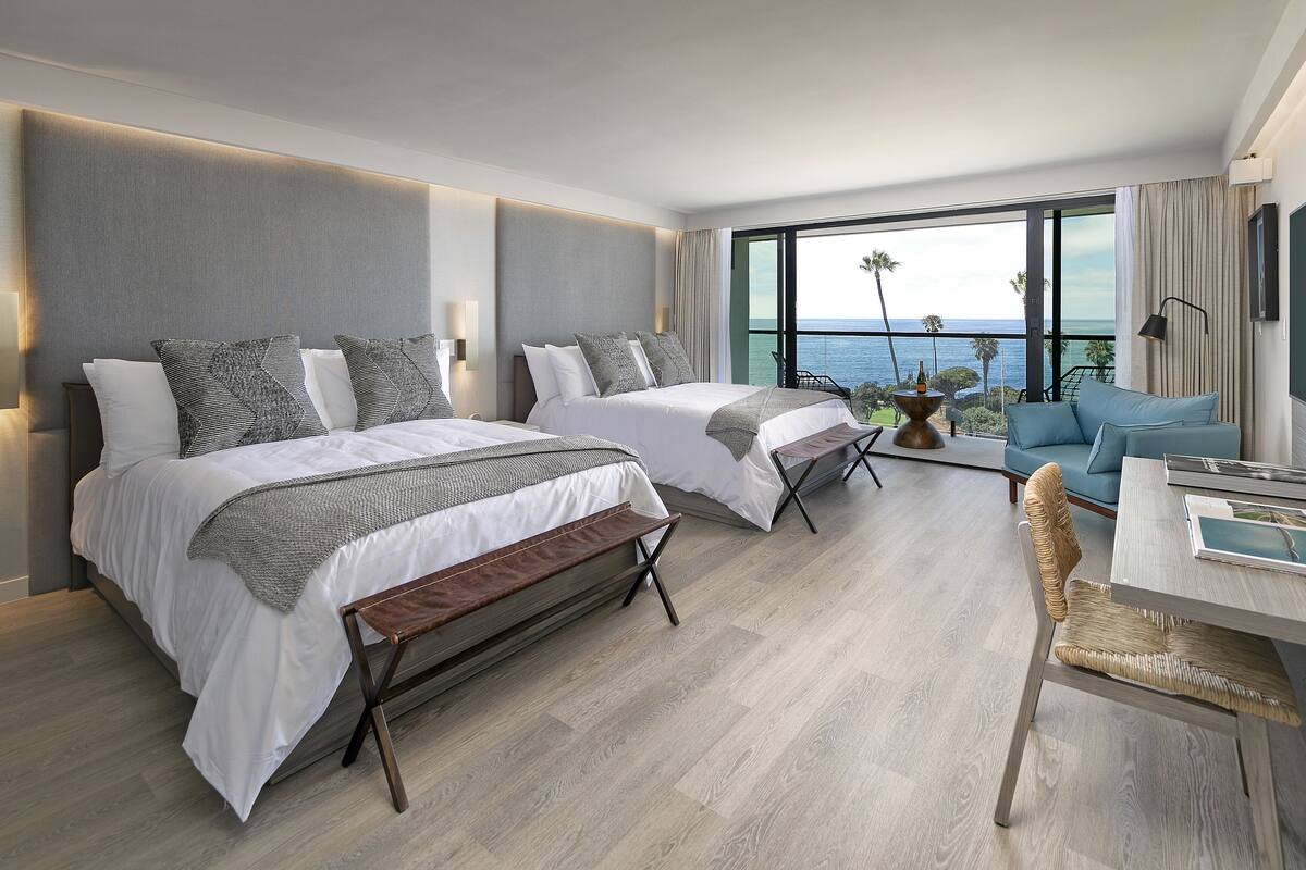 A modern hotel room with two beds, a balcony with a view of the ocean, a seating area, and a dining table.