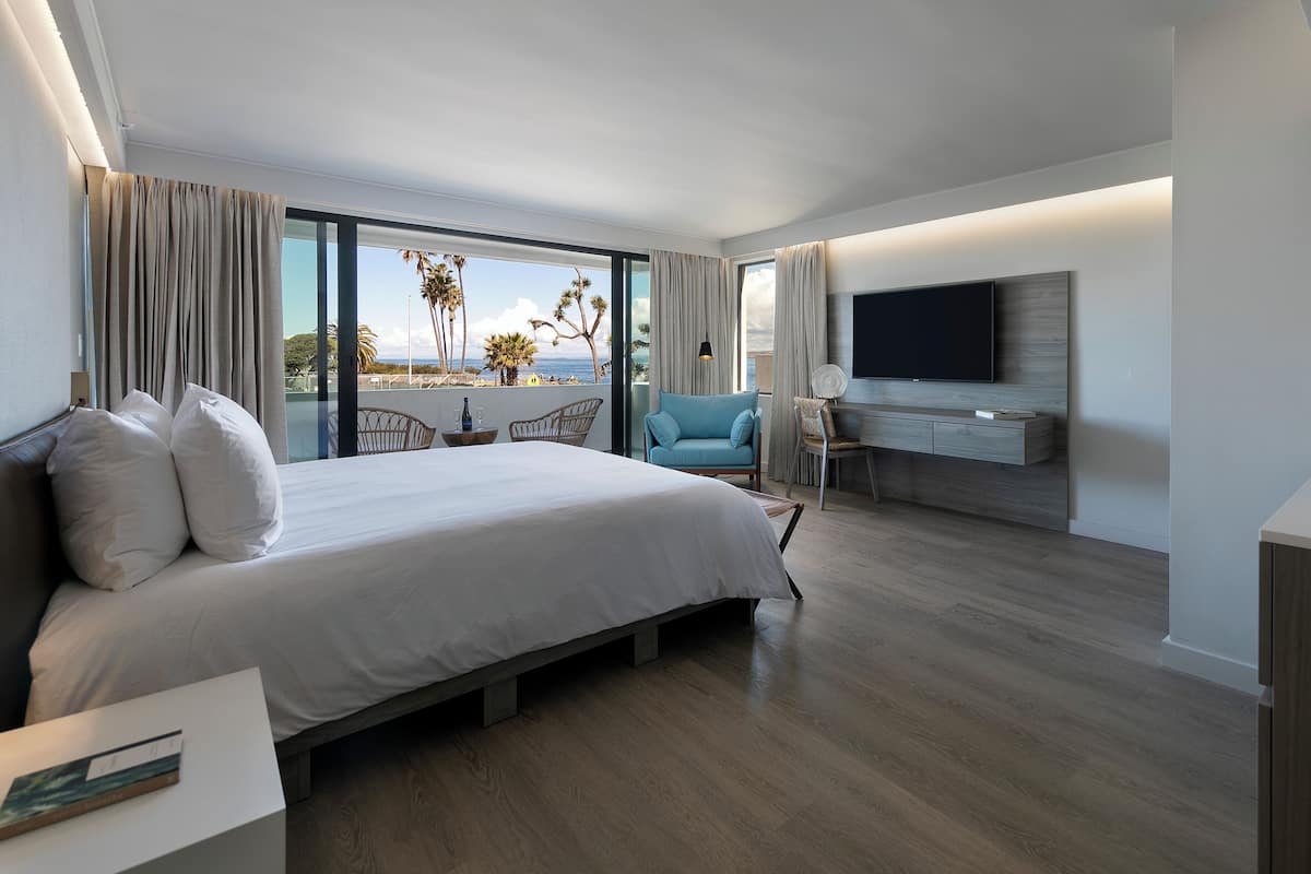 A modern hotel room with a large bed, a desk with a flat-screen TV, and a balcony with a view of palm trees and the sea.
