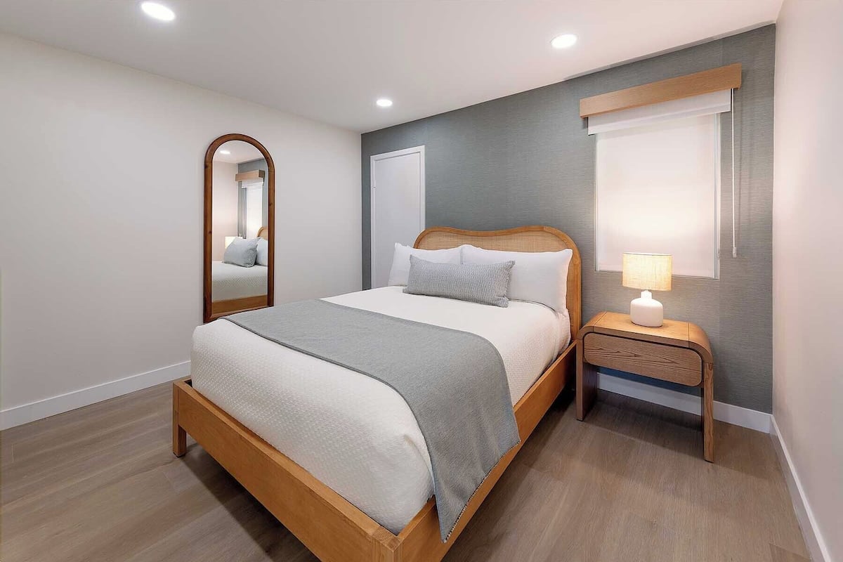 A bedroom with a bed, a nightstand, a lamp, a mirror, and a window.