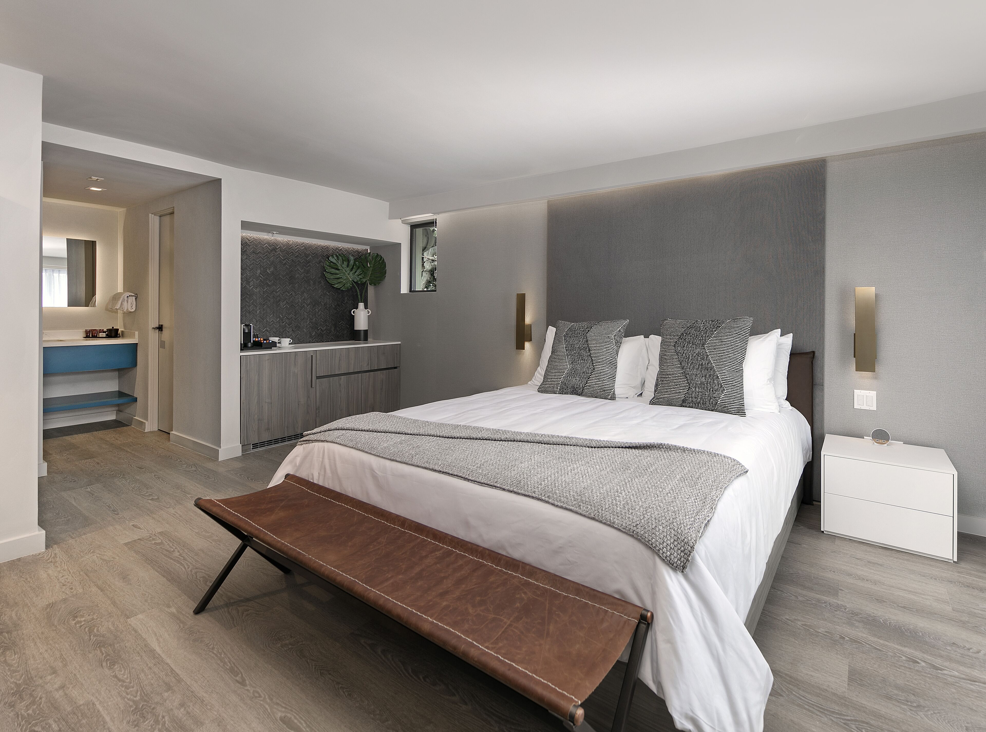 A modern bedroom with a large bed, a bench, a bedside table, and a bathroom visible through an open door.