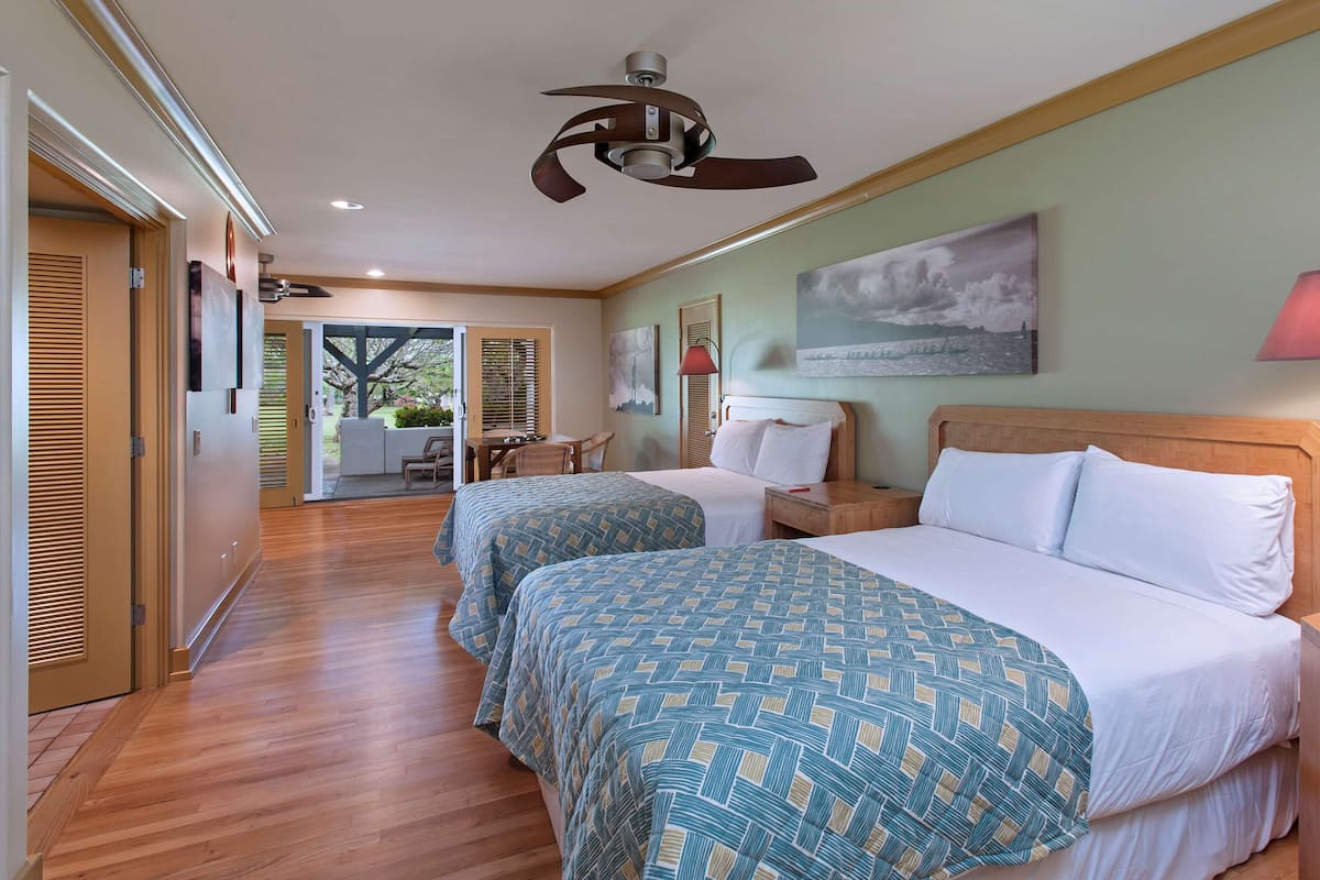 A hotel room with two beds, a ceiling fan, a table with chairs, a lamp, and a painting on the wall.