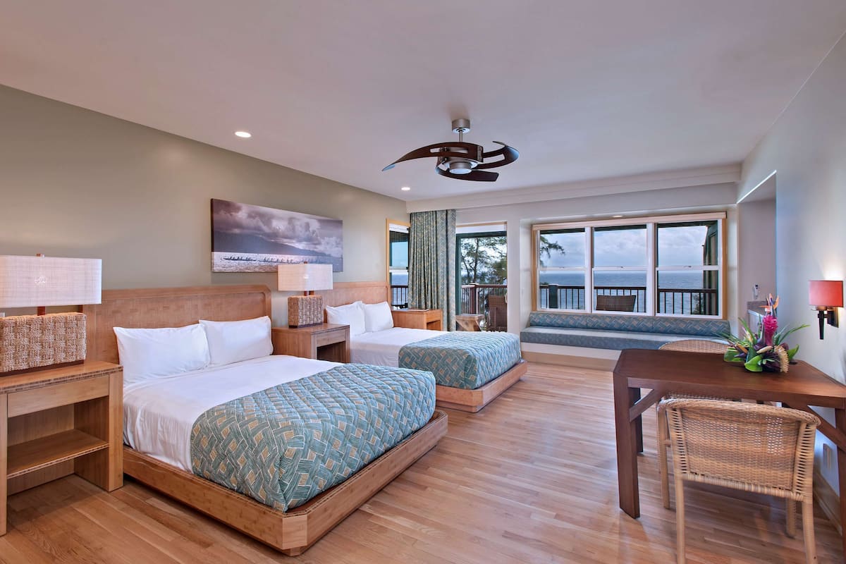 A hotel room with two beds, a wooden floor, a ceiling fan, a large window with a view, and a dining table with chairs.