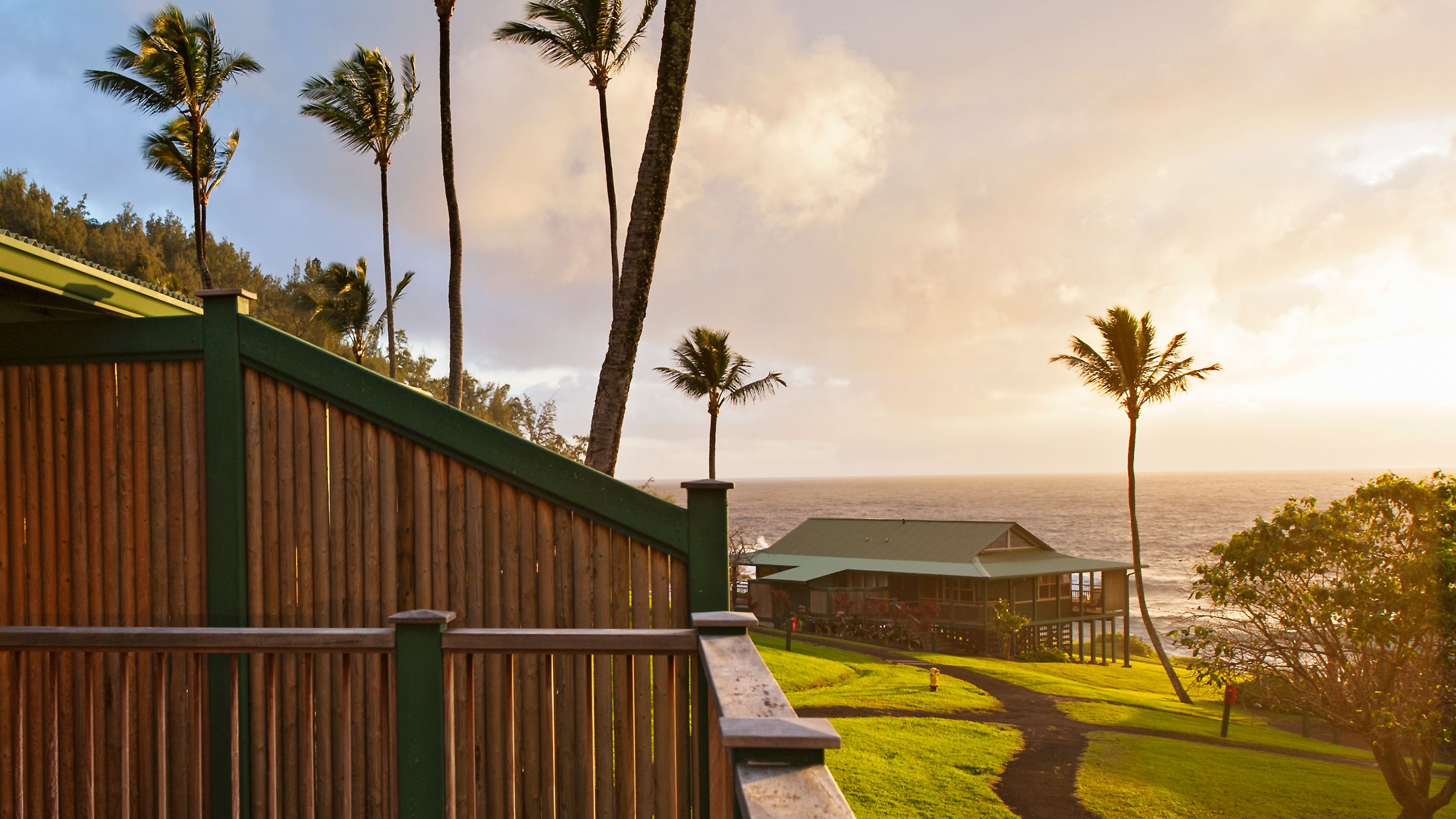 A wooden fence leading to a building with a green roof overlooking the ocean.