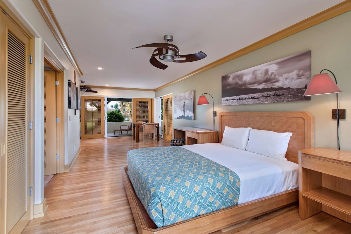A hotel room with a large bed, wooden flooring, a ceiling fan, a desk, a chair, and a view of the outdoors.