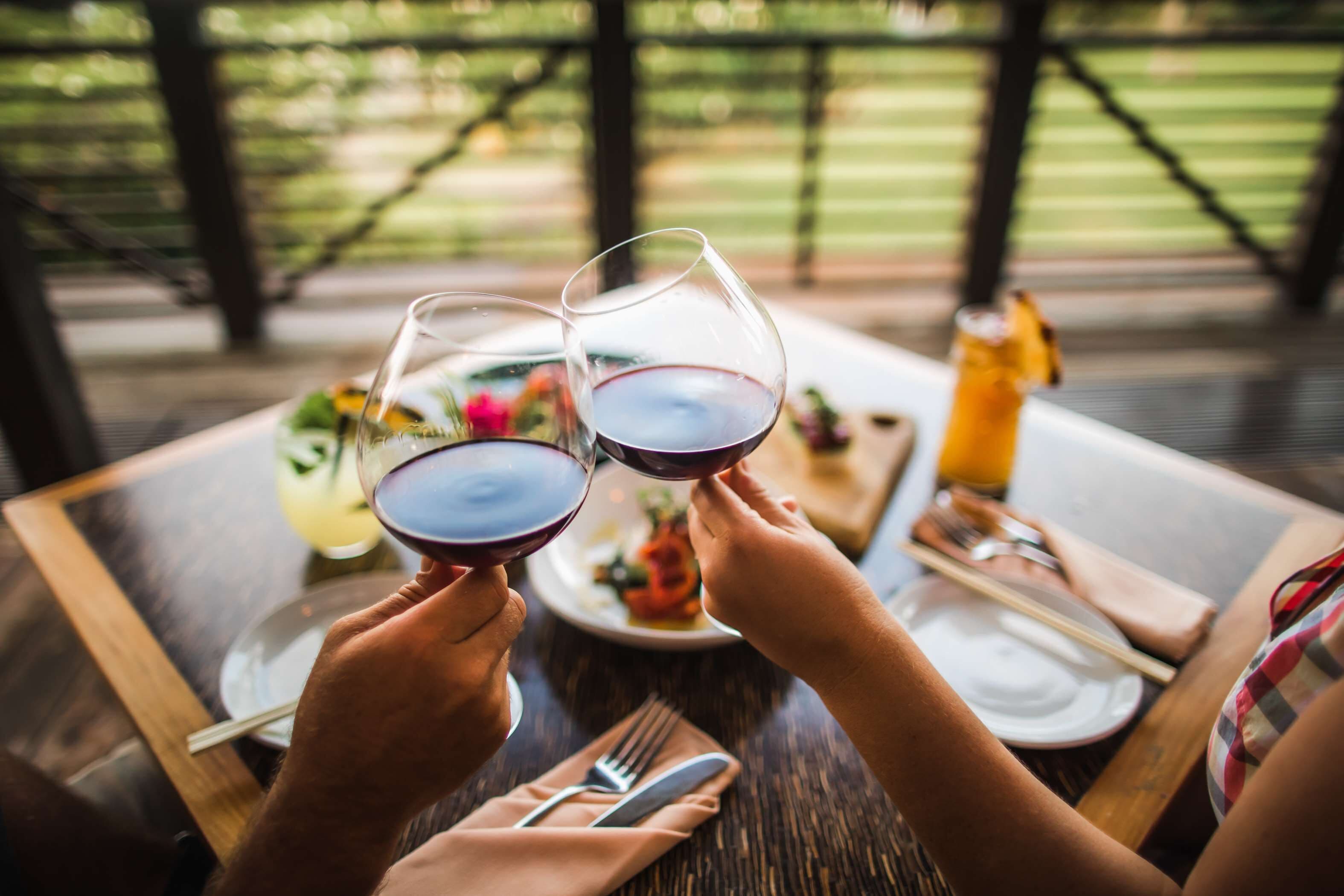 Twee personen die met wijnglazen proosten aan een houten tafel, met een fles wijn, een bord eten en een sapglas.