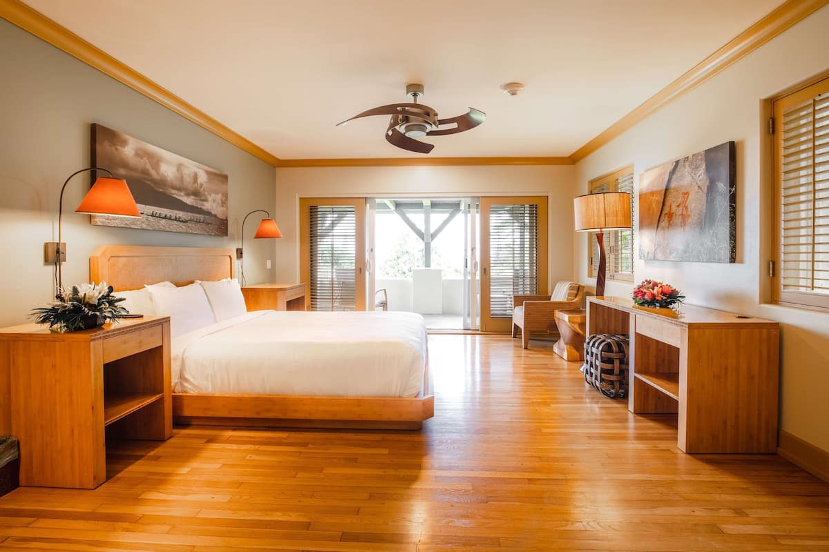 A spacious bedroom with a large bed, wooden flooring, a ceiling fan, and a view through the glass doors.
