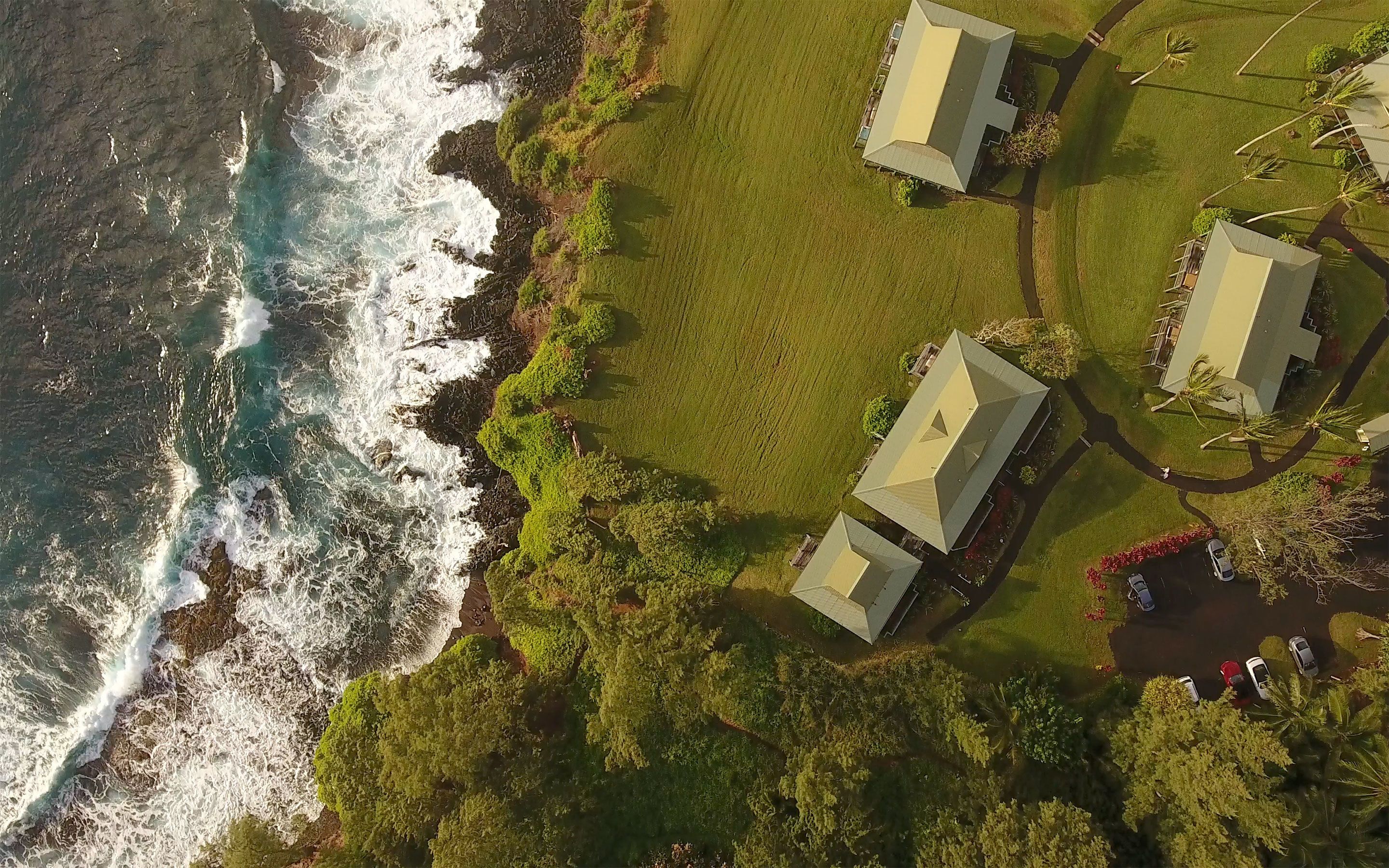 An aerial view of a coastal property with two buildings, a grassy area, and a rocky shoreline with waves.