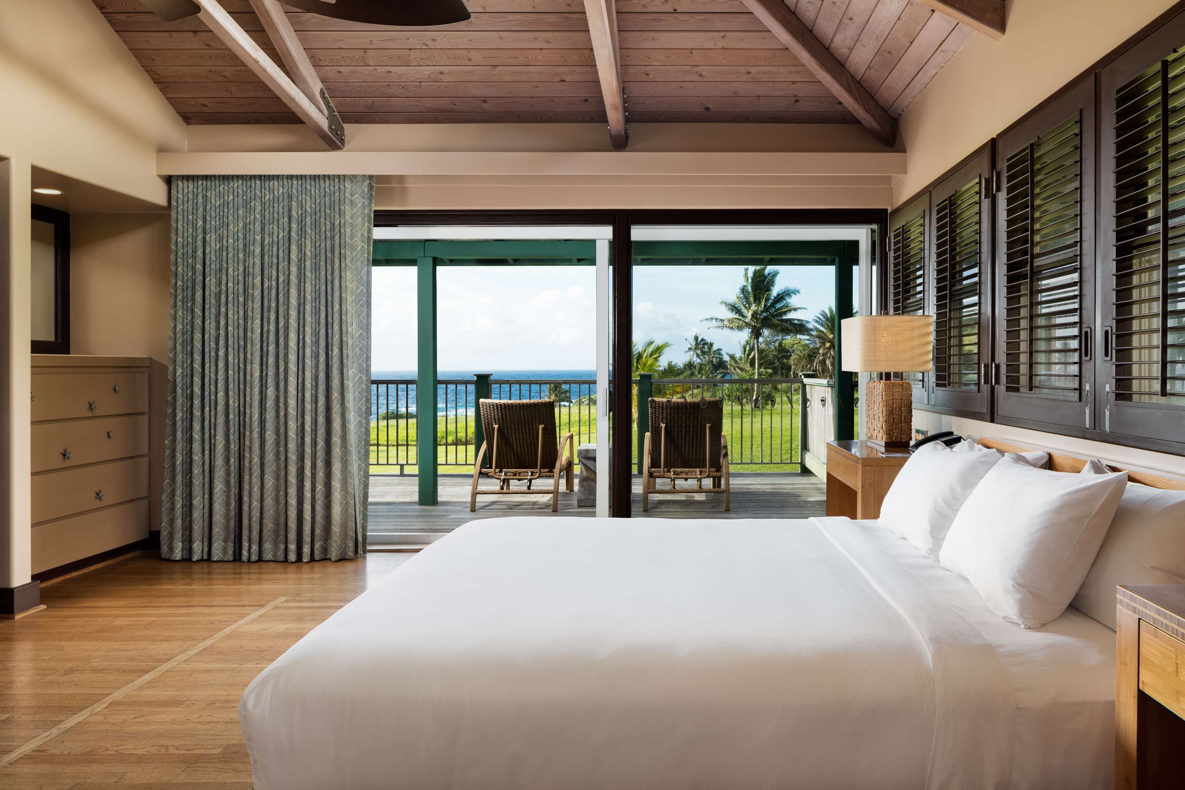 A bedroom with a large bed, wooden ceiling, and a view of the ocean through a sliding glass door.