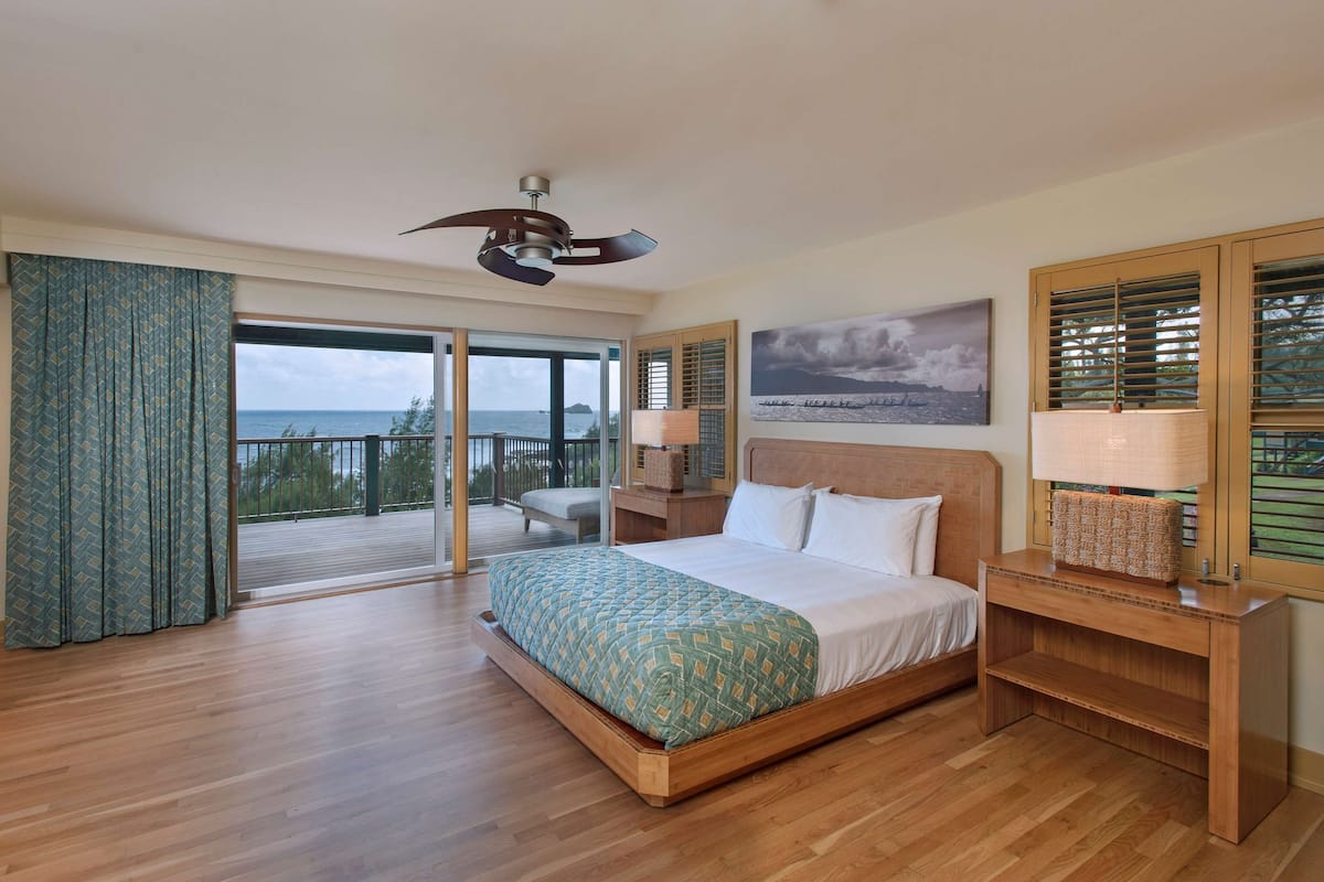 A bedroom with a large bed, a ceiling fan, a nightstand, a lamp, and a view of the ocean through a sliding glass door.