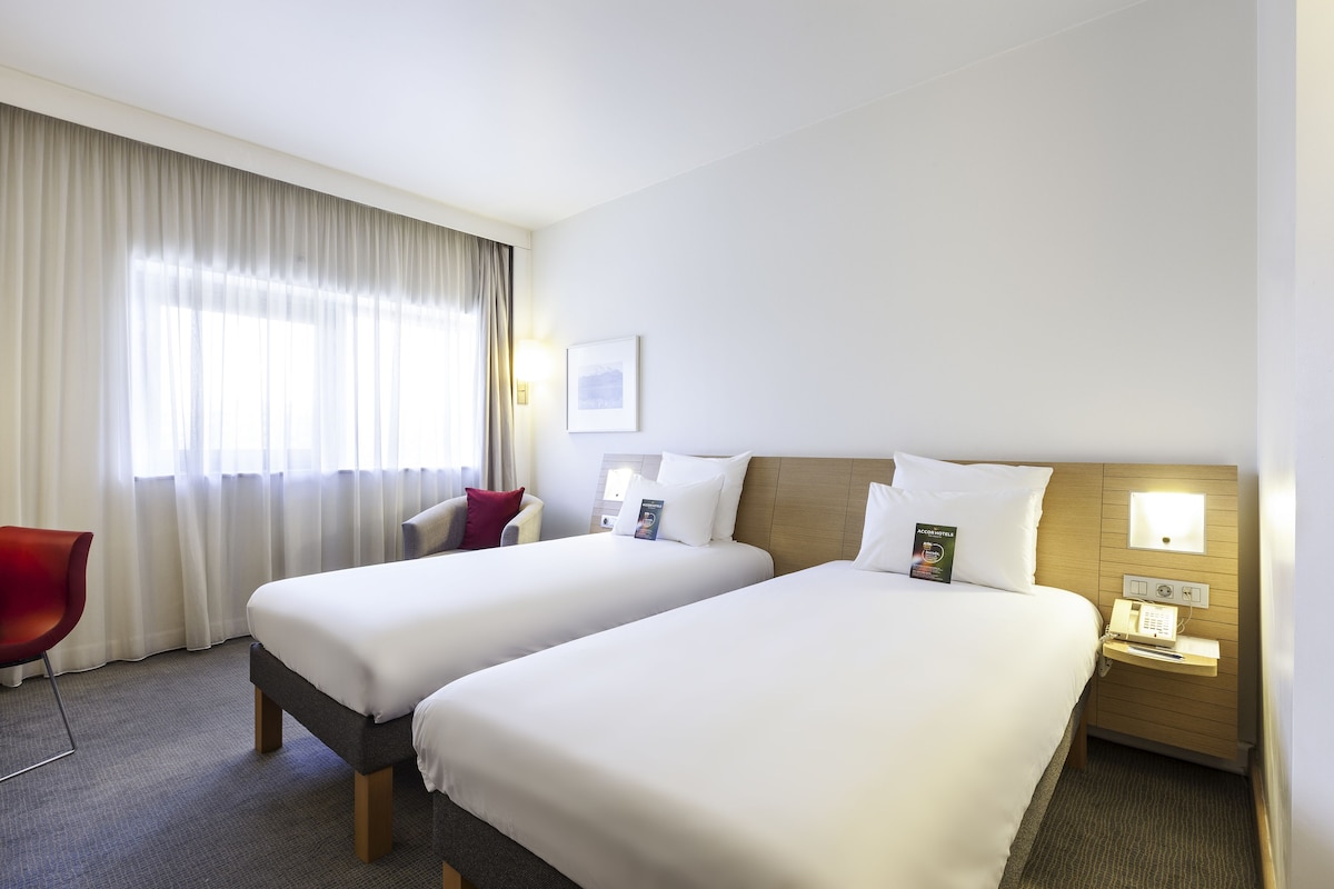 Habitació d'hotel amb dos llits, una cadira vermella, una taula de nit amb un telèfon i una finestra amb cortines.