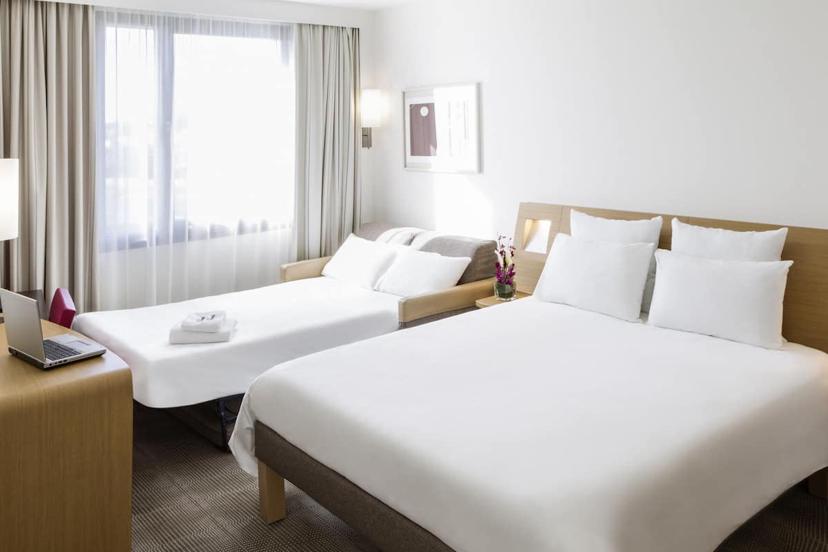 Ein Hotelzimmer mit zwei Betten, einem Schreibtisch mit einem Laptop, einem Nachttisch und einem Fenster mit Vorhängen.